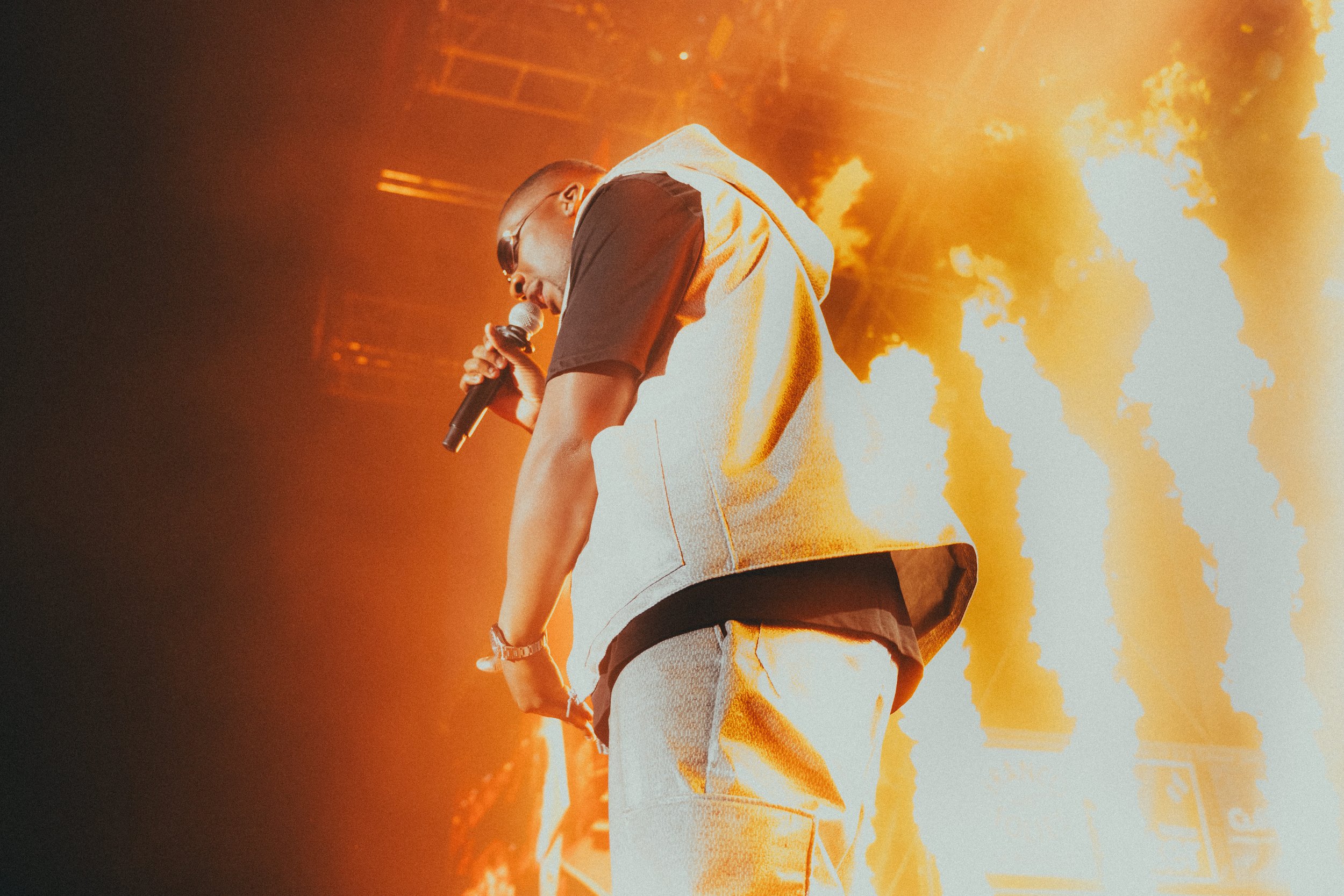 Un performer portant des vêtements élégants sur scène, tenant un microphone, avec des effets de feu et de lumière orange en arrière-plan.