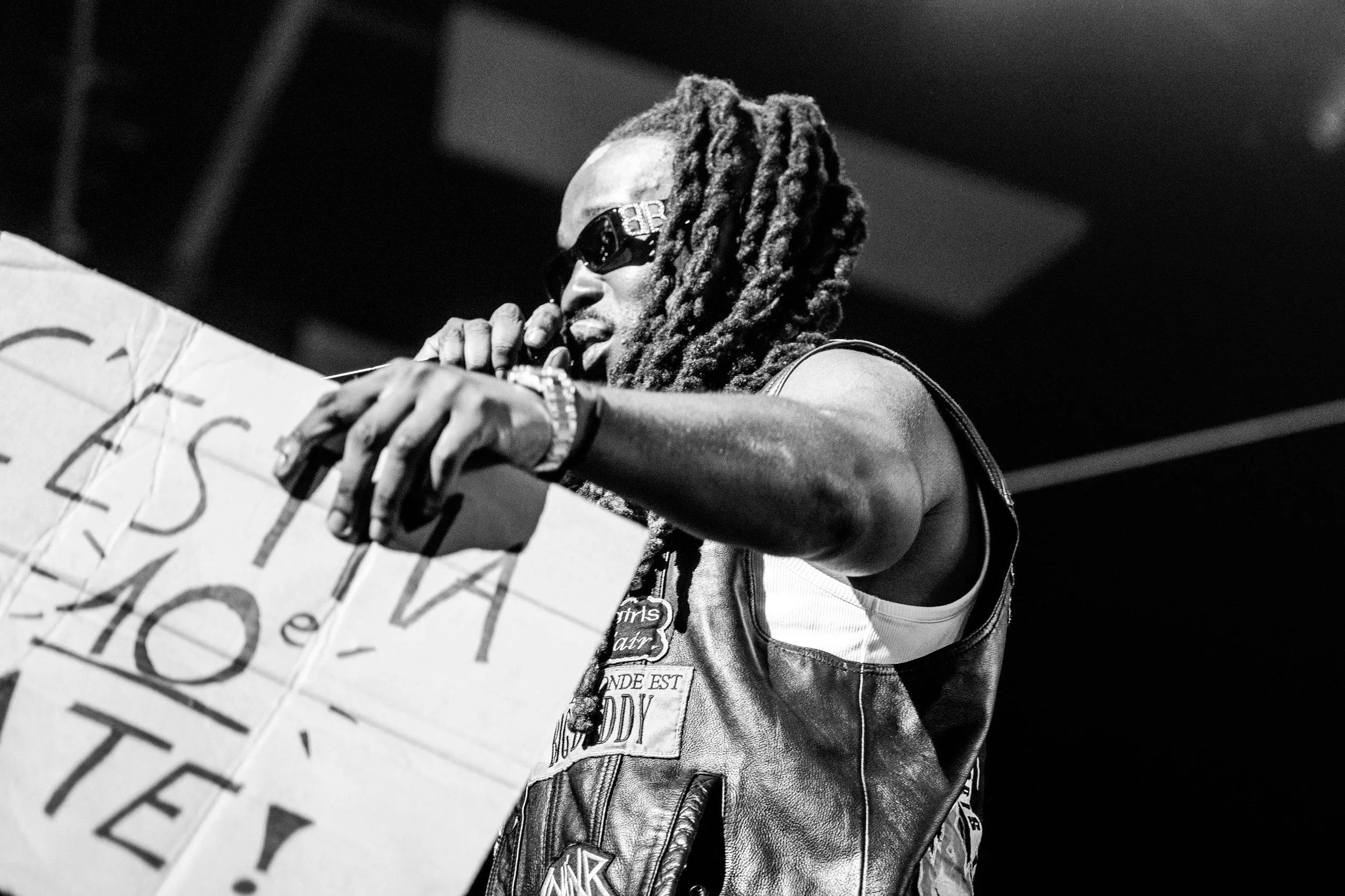 Un homme avec des dreadlocks portant des lunettes de soleil et un gilet en cuir noir tient un panneau en papier avec des lettres, lors d'une performance ou d'une manifestation.