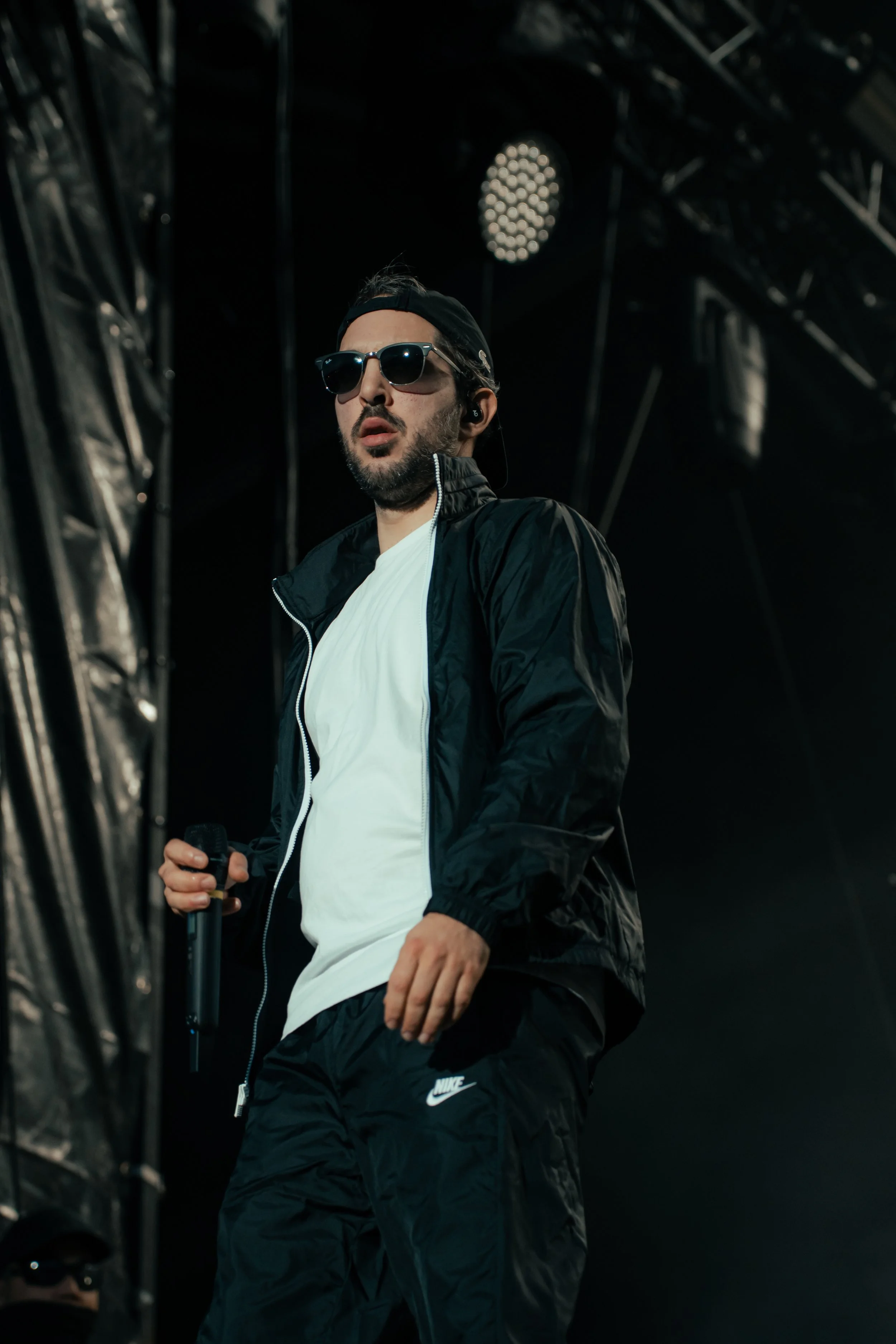 Un homme portant des lunettes de soleil, un casque, un pantalon de sport Nike et une veste noire, tenant un micro sur scène.