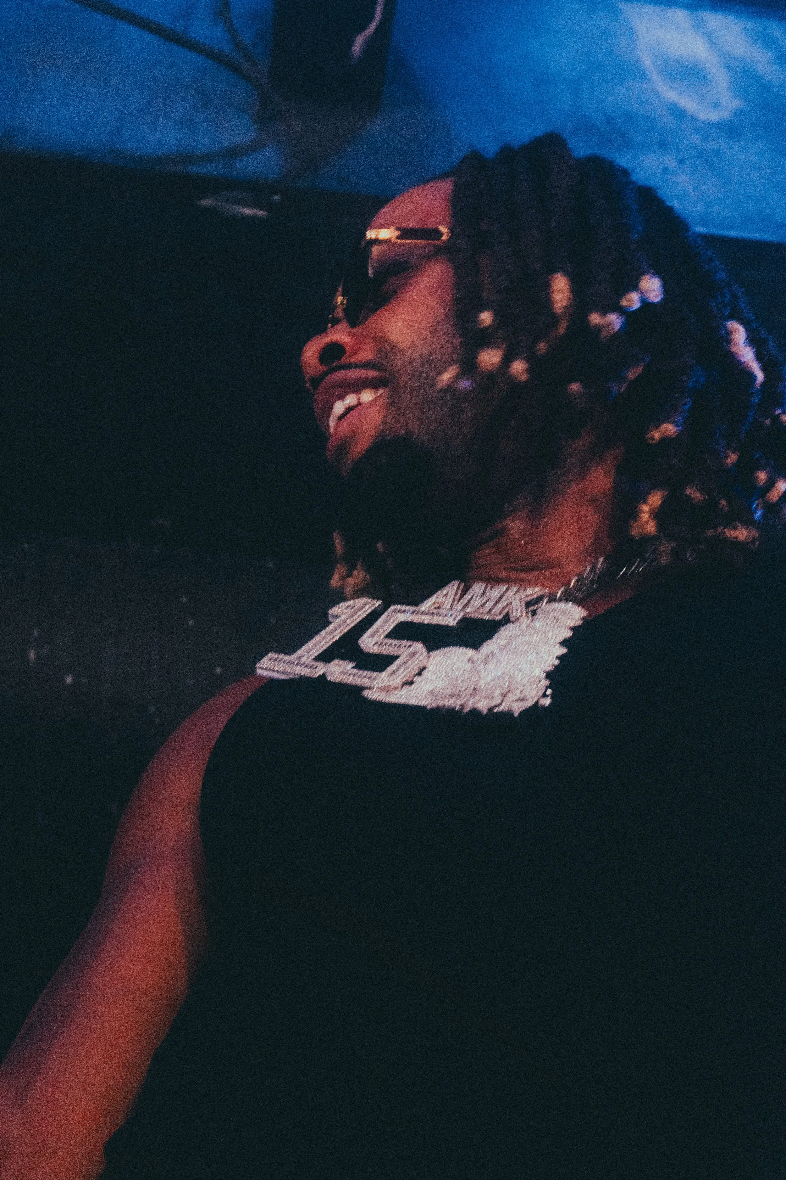 Homme avec dreadlocks portant des lunettes de soleil, un collier en forme de « 1501 » en diamants, souriant en regardant vers la gauche, dans un environnement sombre et coloré.