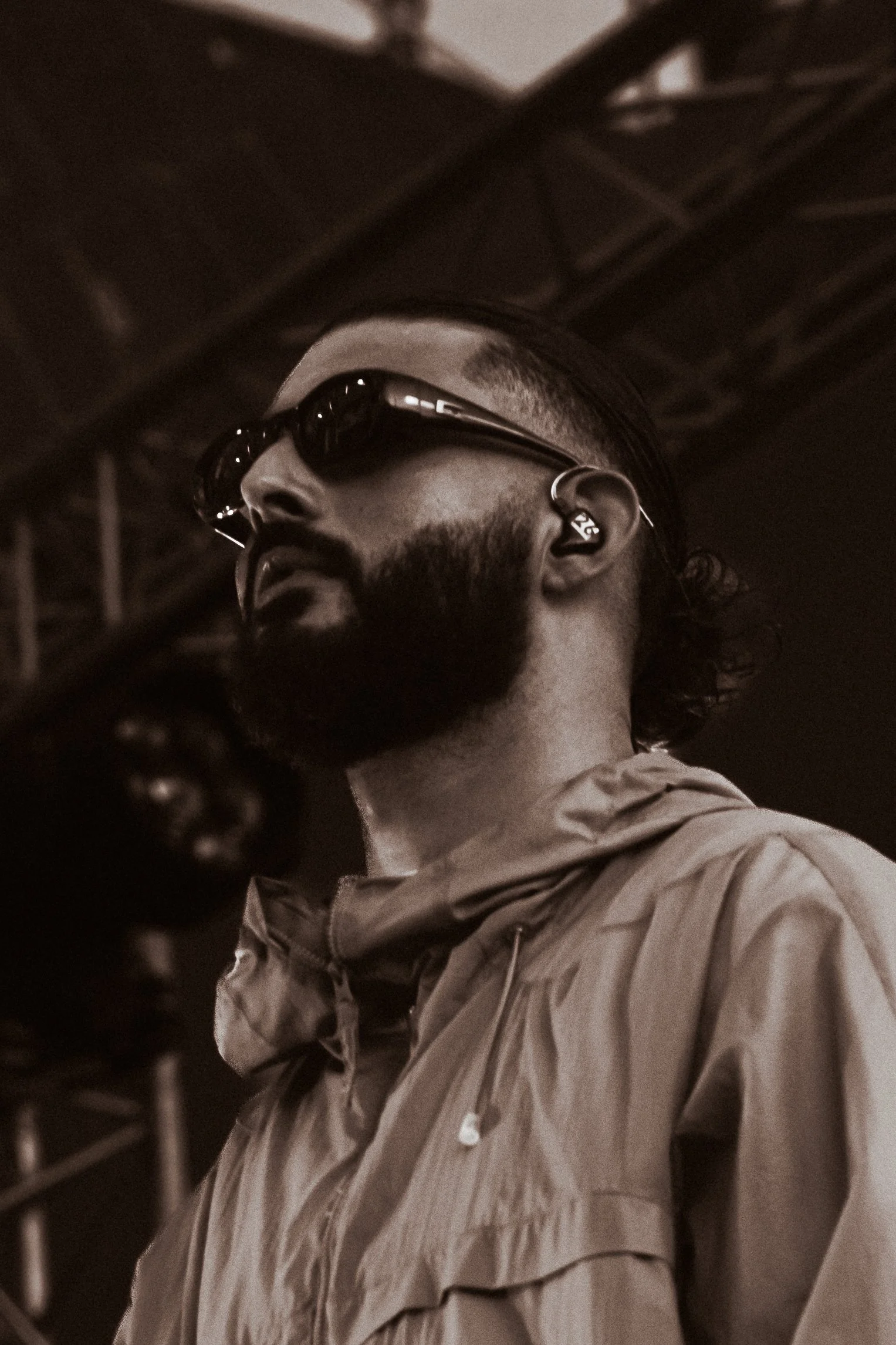 Un homme avec une barbe portant des lunettes de soleil foncées, une oreillette et une veste en tissu, photographié de profil dans une ambiance sombre.