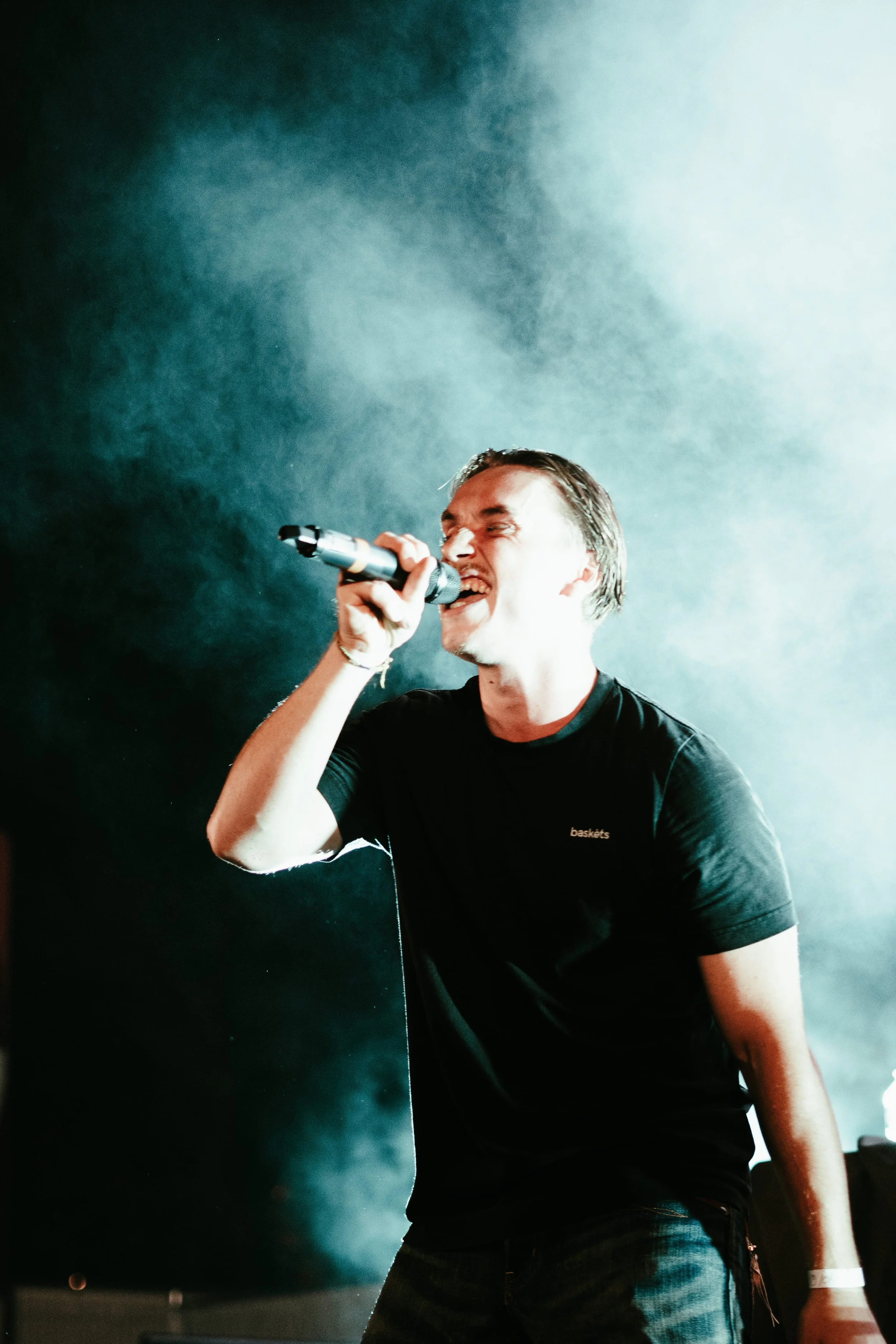 Un homme chante en tenant un microphone dans un environnement sombre illuminé par des lumières, avec de la fumée en arrière-plan.