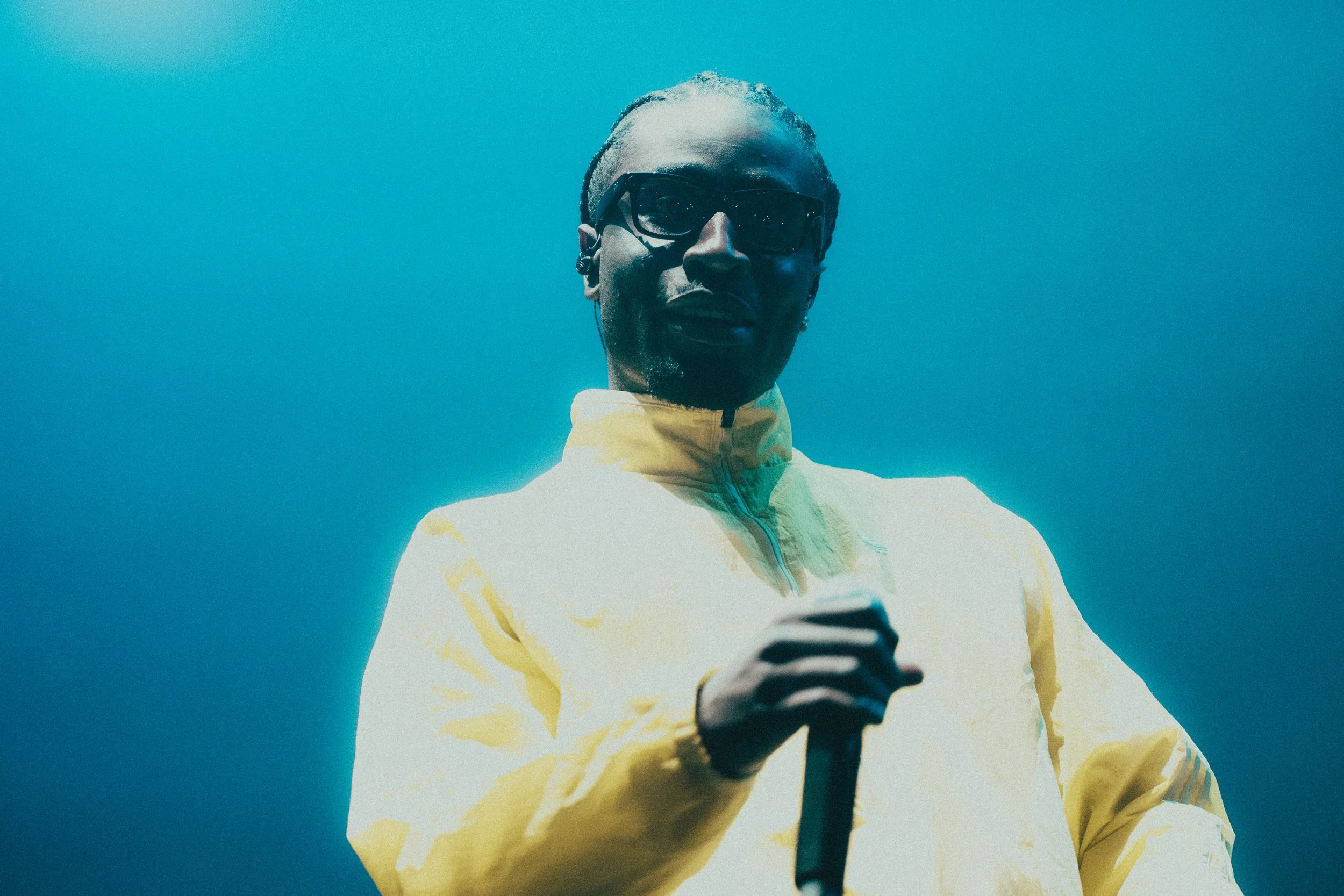 Un homme porte des lunettes de soleil et une veste jaune, tenant un microphone, avec un ciel bleu en arrière-plan.