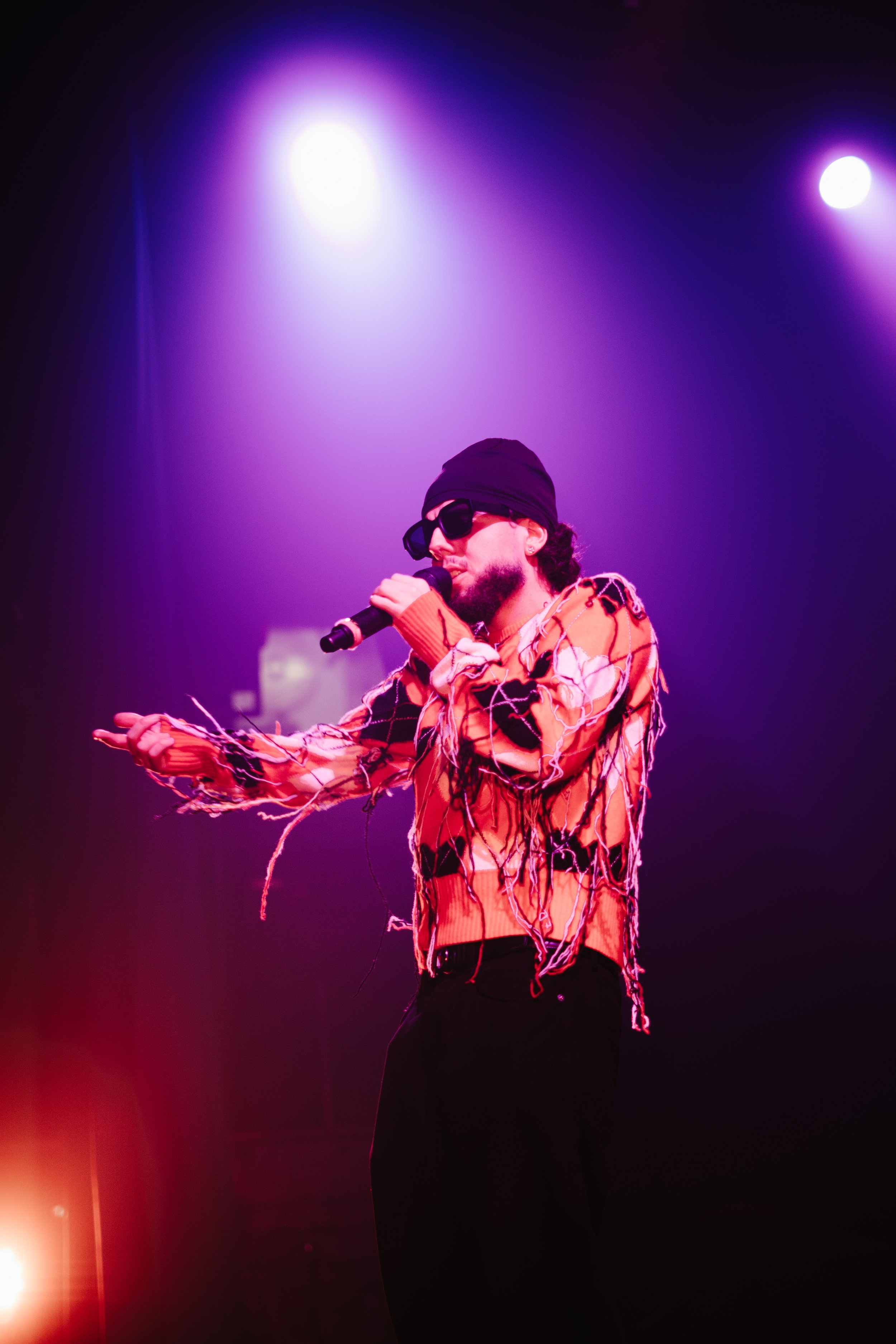 Un homme avec un béret, des lunettes de soleil, et un sweat à motifs colorés, chante avec un micro sur scène sous des lumières violettes et roses.