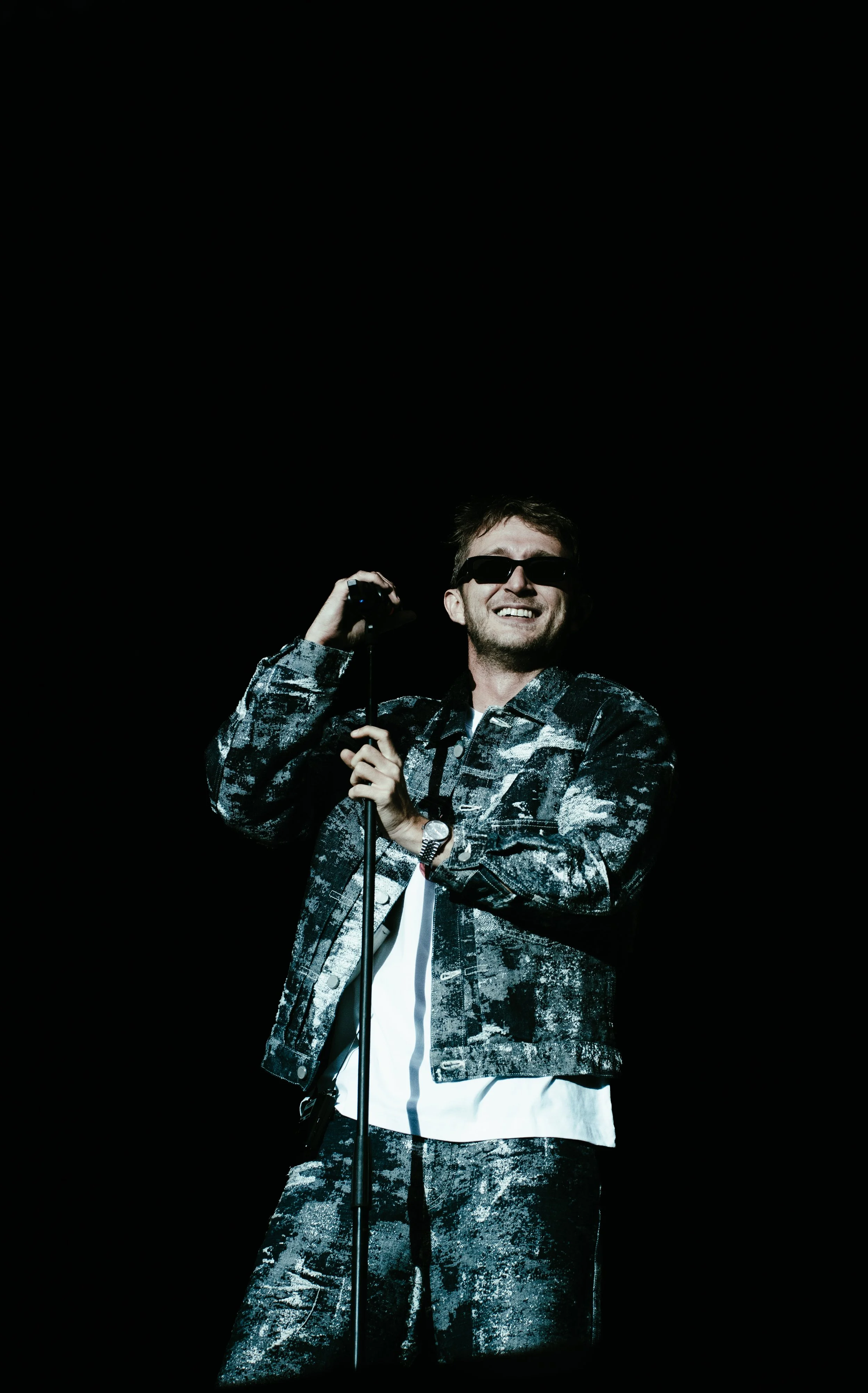 Un homme portant des lunettes de soleil, une veste camouflage, et une montre, souriant, tenant un microphone, dans un fond noir.