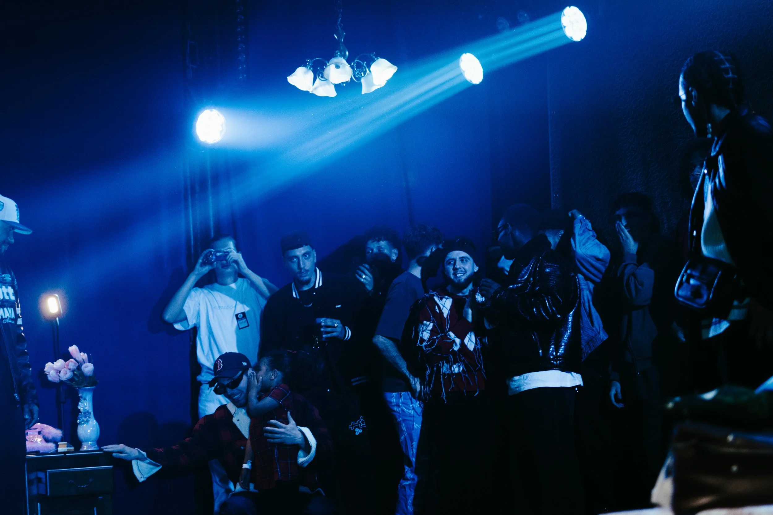 Groupe de personnes dans une pièce sombre avec éclairage bleu, certains discutant, d'autres prenant des photos ou regardant. Un lustre blanc au plafond et une table avec des fleurs