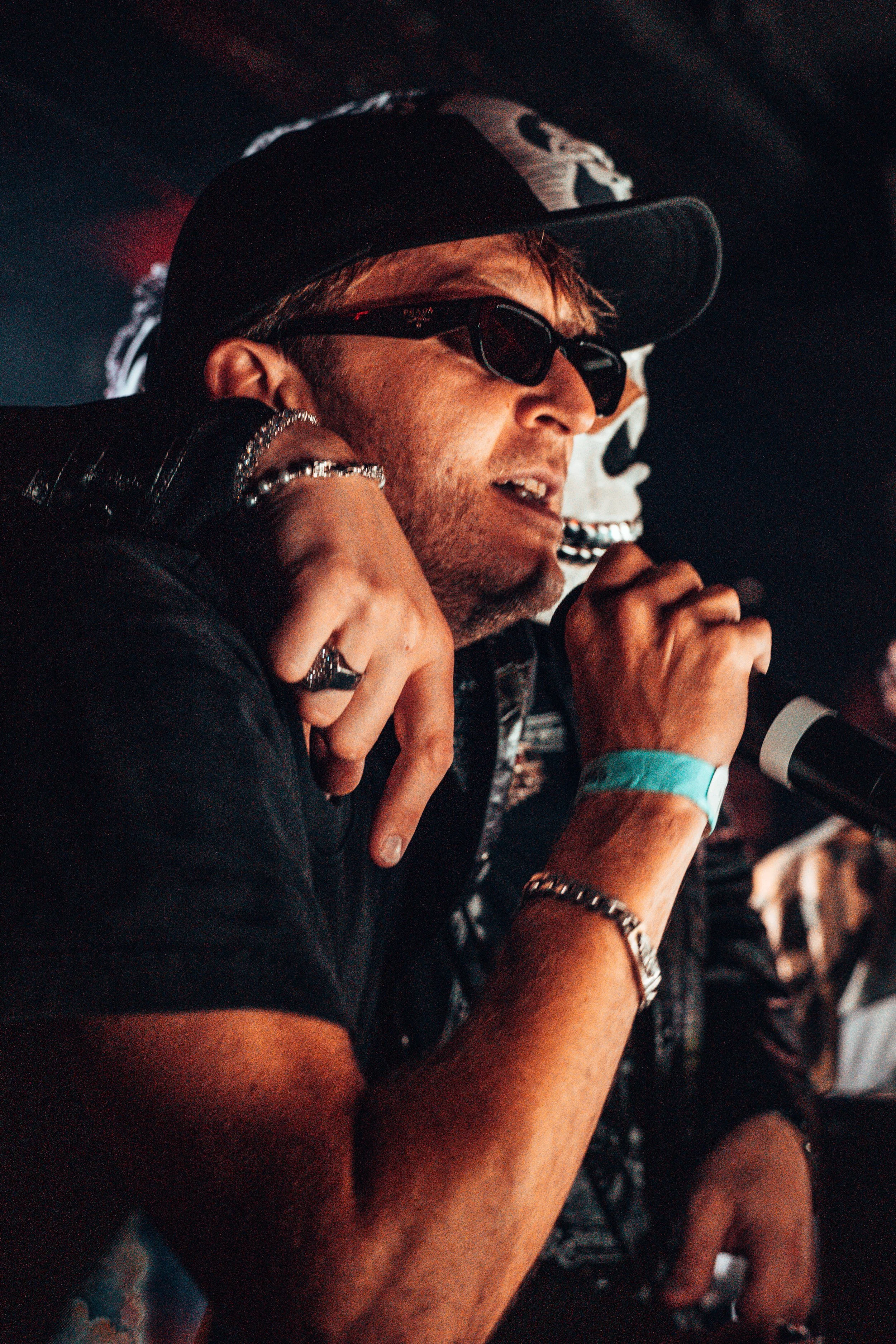 Homme portant des lunettes de soleil, une casquette, tenant un micro, avec des bijoux et un bracelet en silicone bleu, en train de parler ou chanter.