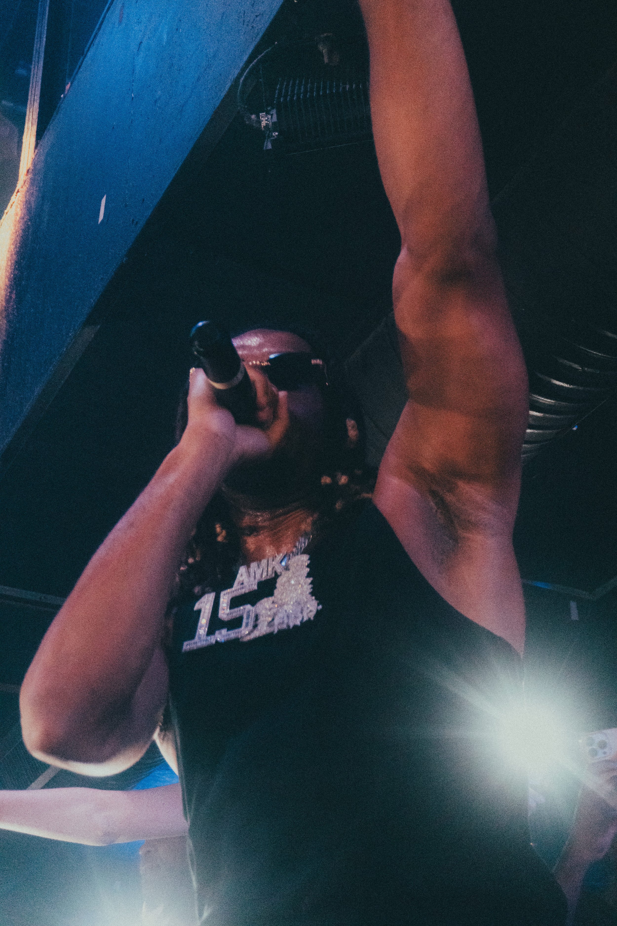 Une personne portant un t-shirt noir avec un motif de texte et un nombre 15, vue de dessus, tenant un micro dans sa main, dans un environnement sombre avec quelques lumières