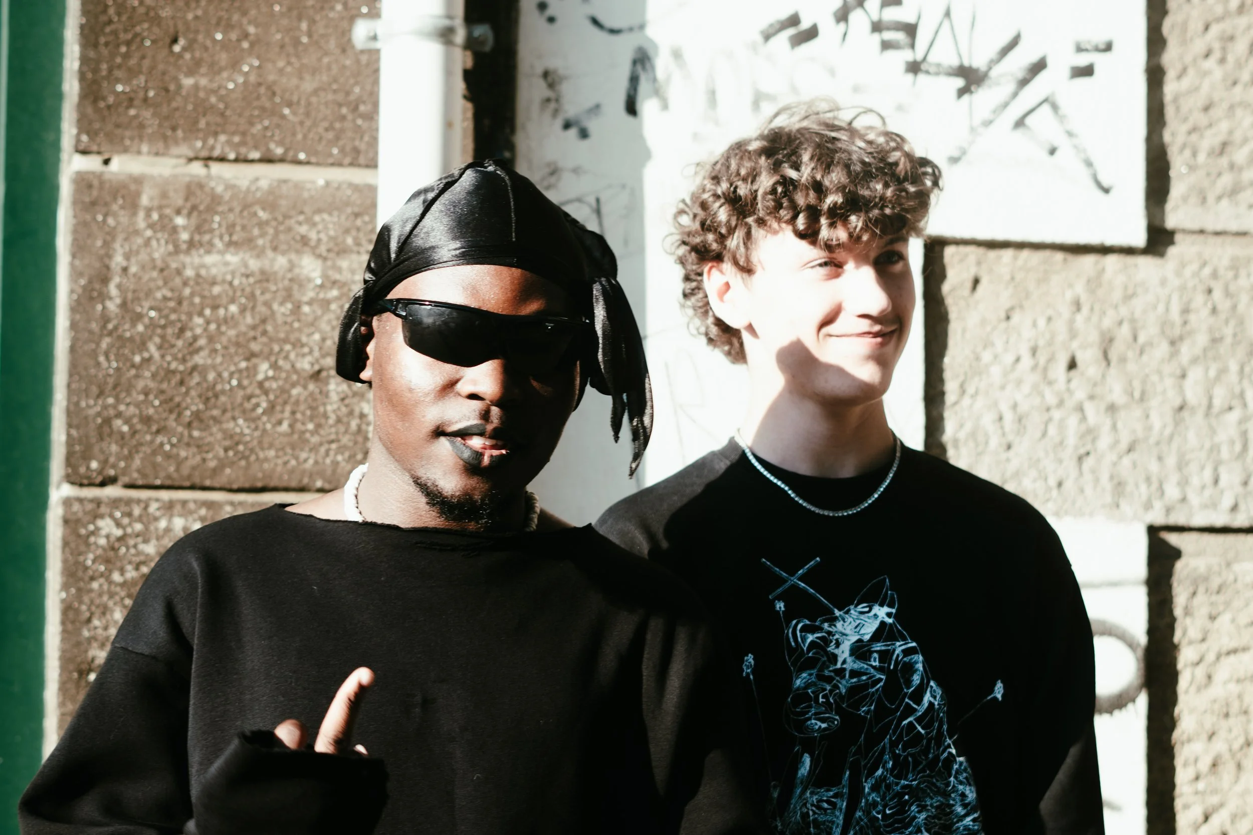 Deux jeunes hommes posant devant un mur en pierre et un panneau blanc. L'un porte des lunettes de soleil noires, un bonnet noir, une chaîne et un sweat noir. L'autre a des cheveux bouclés, porte un T-shirt noir avec un design graphique et une chaîne,