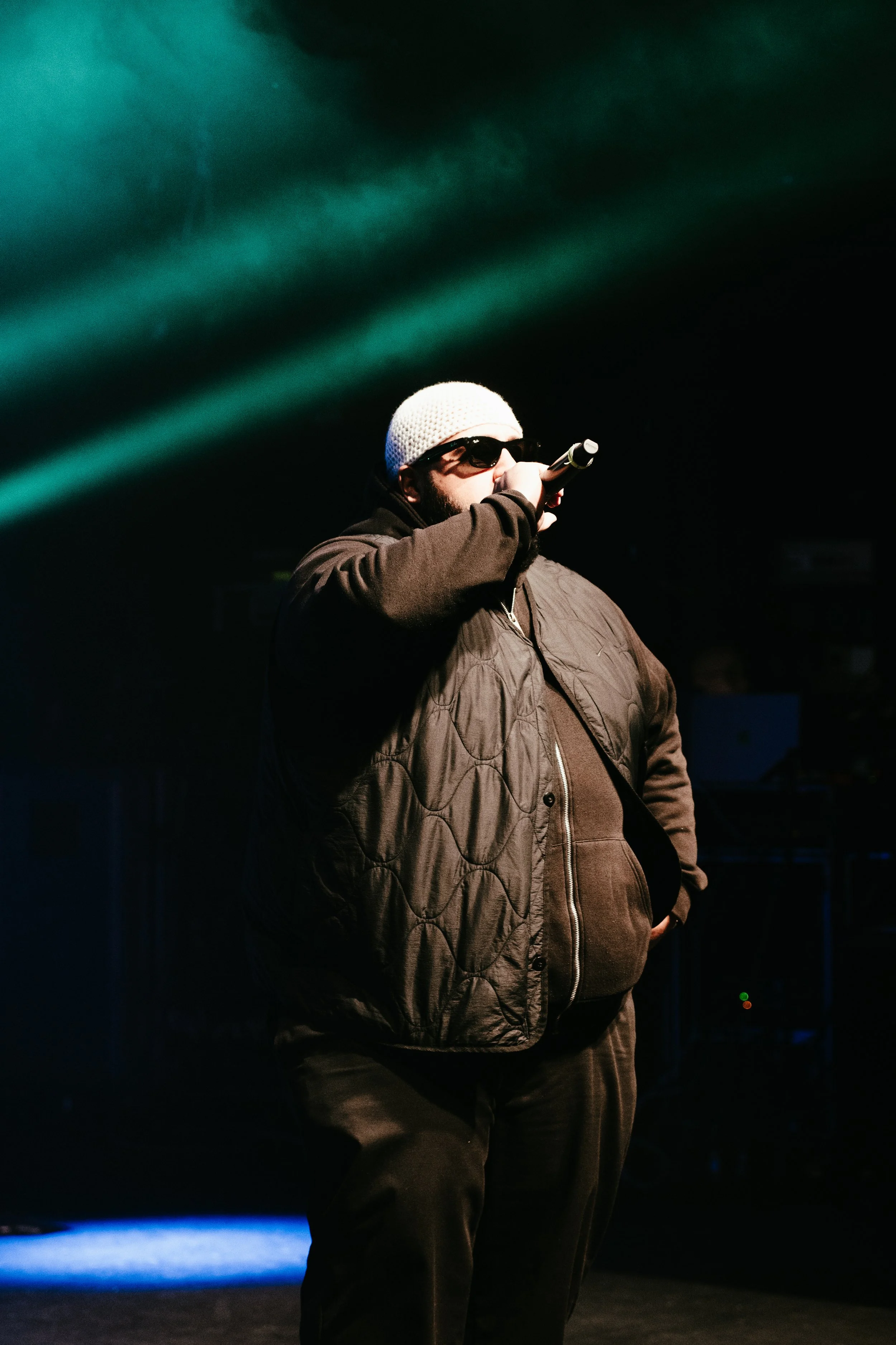 Un homme musclé portant des lunettes de soleil, un bonnet blanc, et une veste noire, tenant un microphone sur scène avec un fond sombre et des faisceaux de lumière verte.