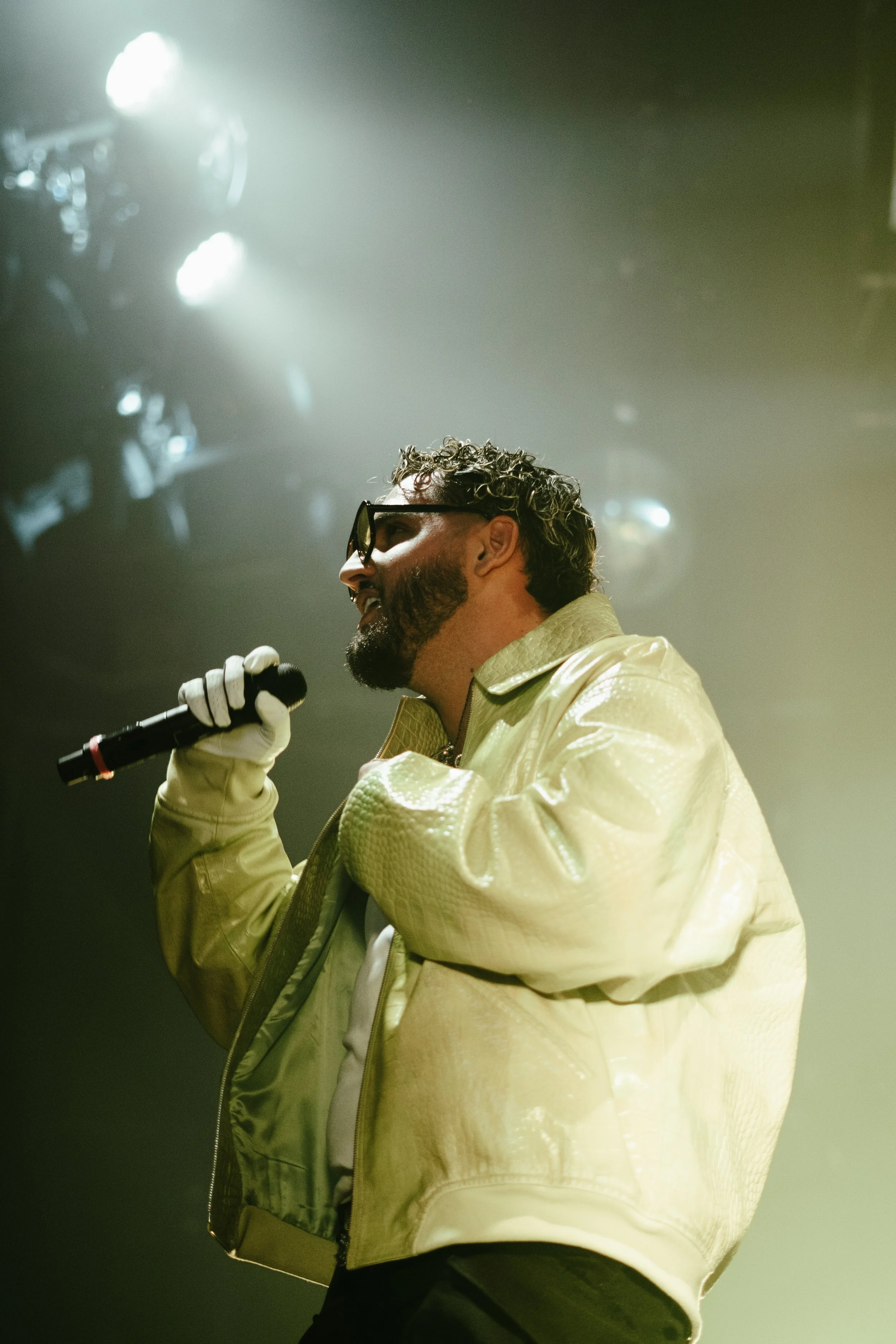 Un homme portant des lunettes de soleil et un blouson jaune clair tient un microphone lors d'une performance sur scène, avec des lumières brillantes en arrière-plan.