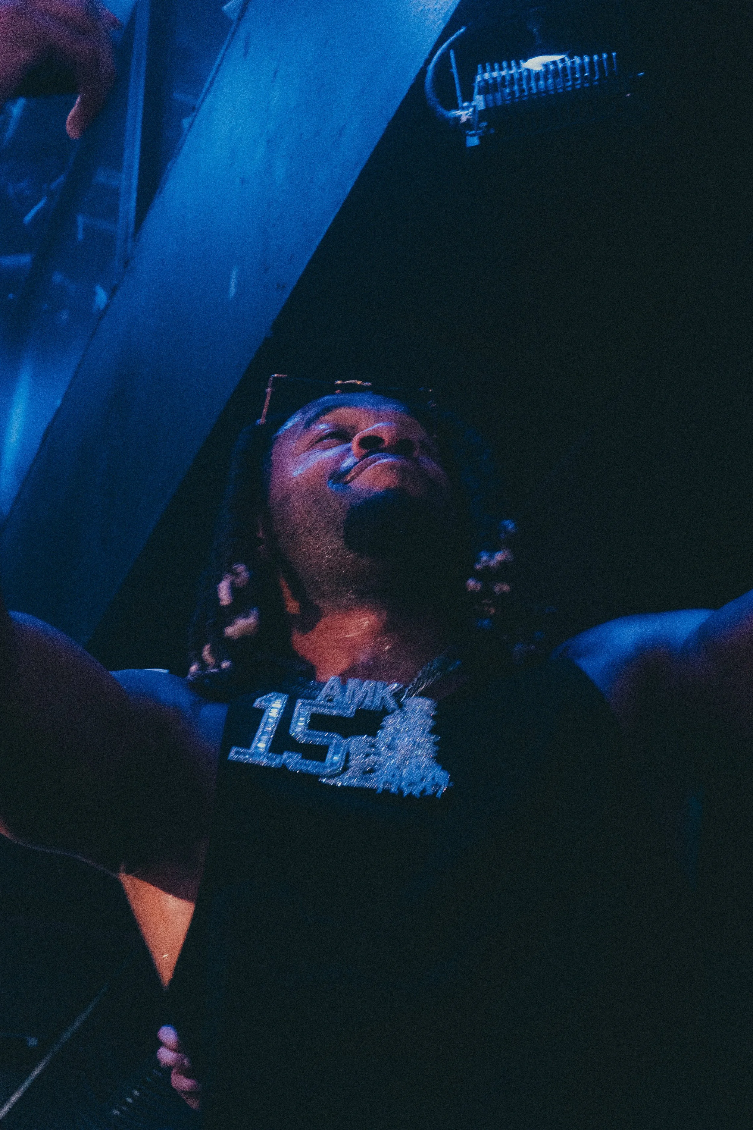 Une personne avec une chaîne en or portant un collier avec la mention "15" prend une selfie dans une pièce sombre, éclairée par une lumière bleue.