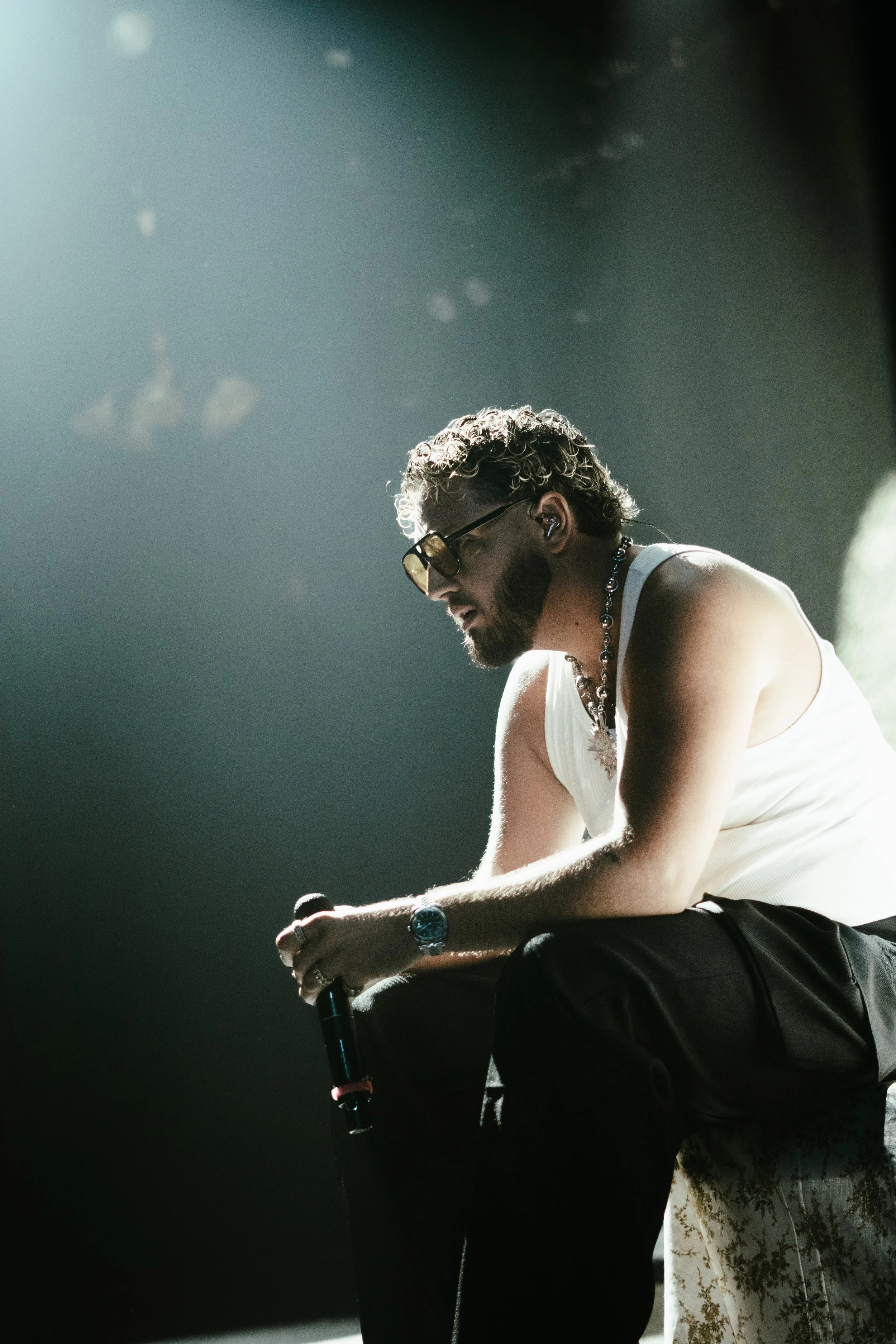 Un homme assis sur une scène, tenant un microphone, portant des lunettes de soleil, un collier, un bracelet, un t-shirt blanc et un pantalon noir.