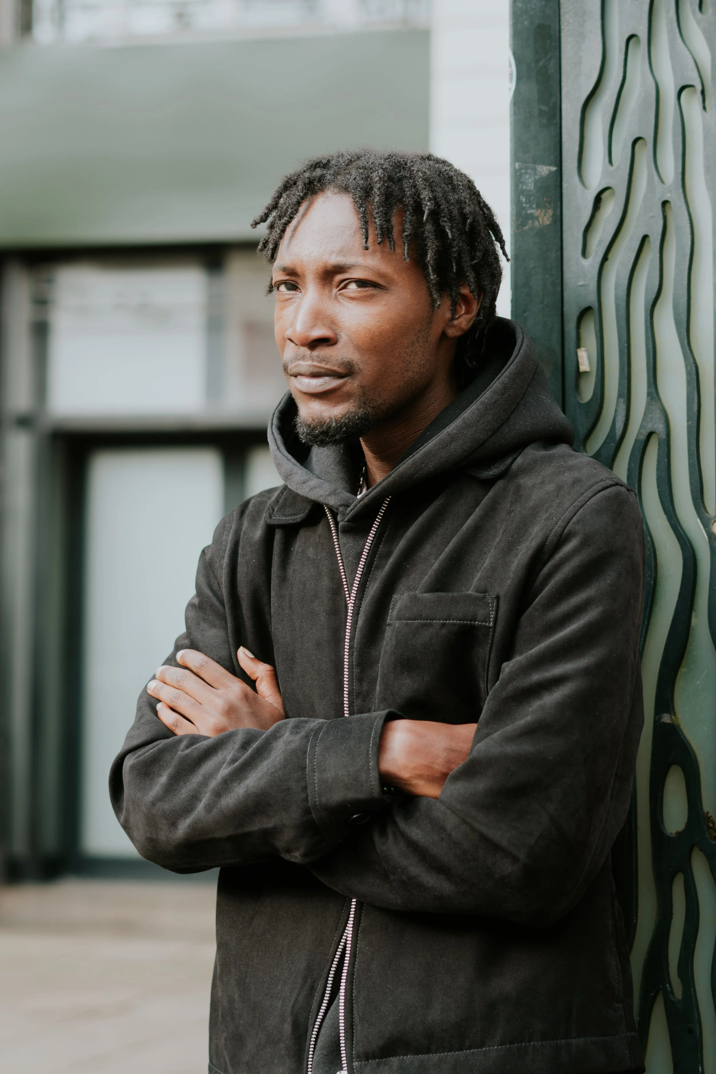 Homme debout contre un mur en métal, bras croisés, regardant vers l'objectif, portant une veste noire et un sweat à capuche