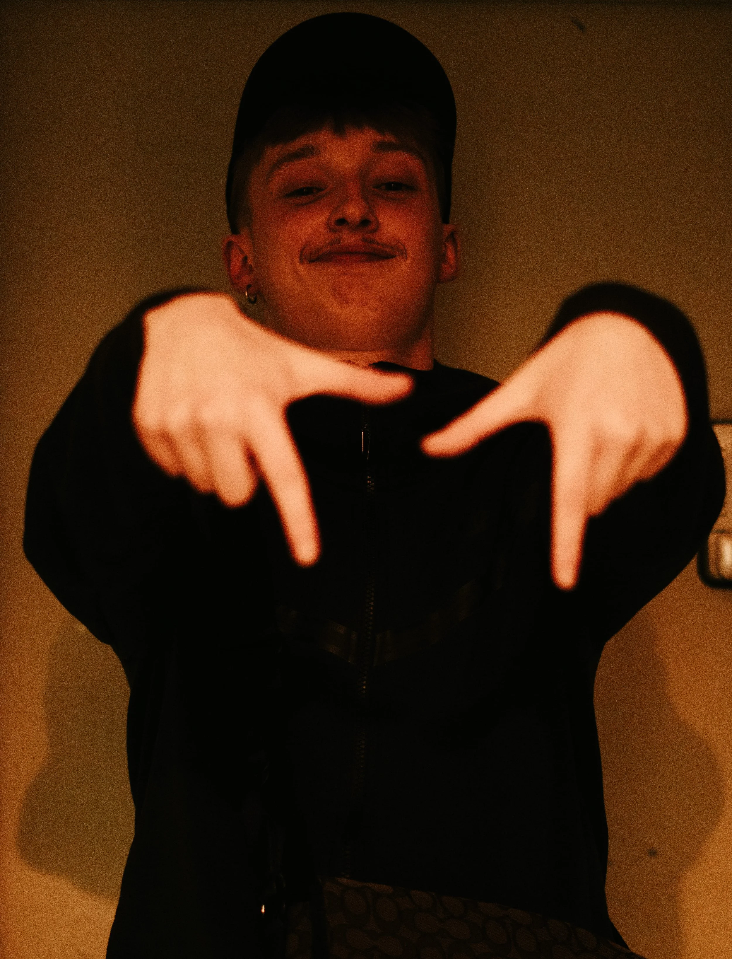 Jeune homme portant une casquette, souriant, pointant vers le bas avec les deux mains, dans une ambiance sombre.