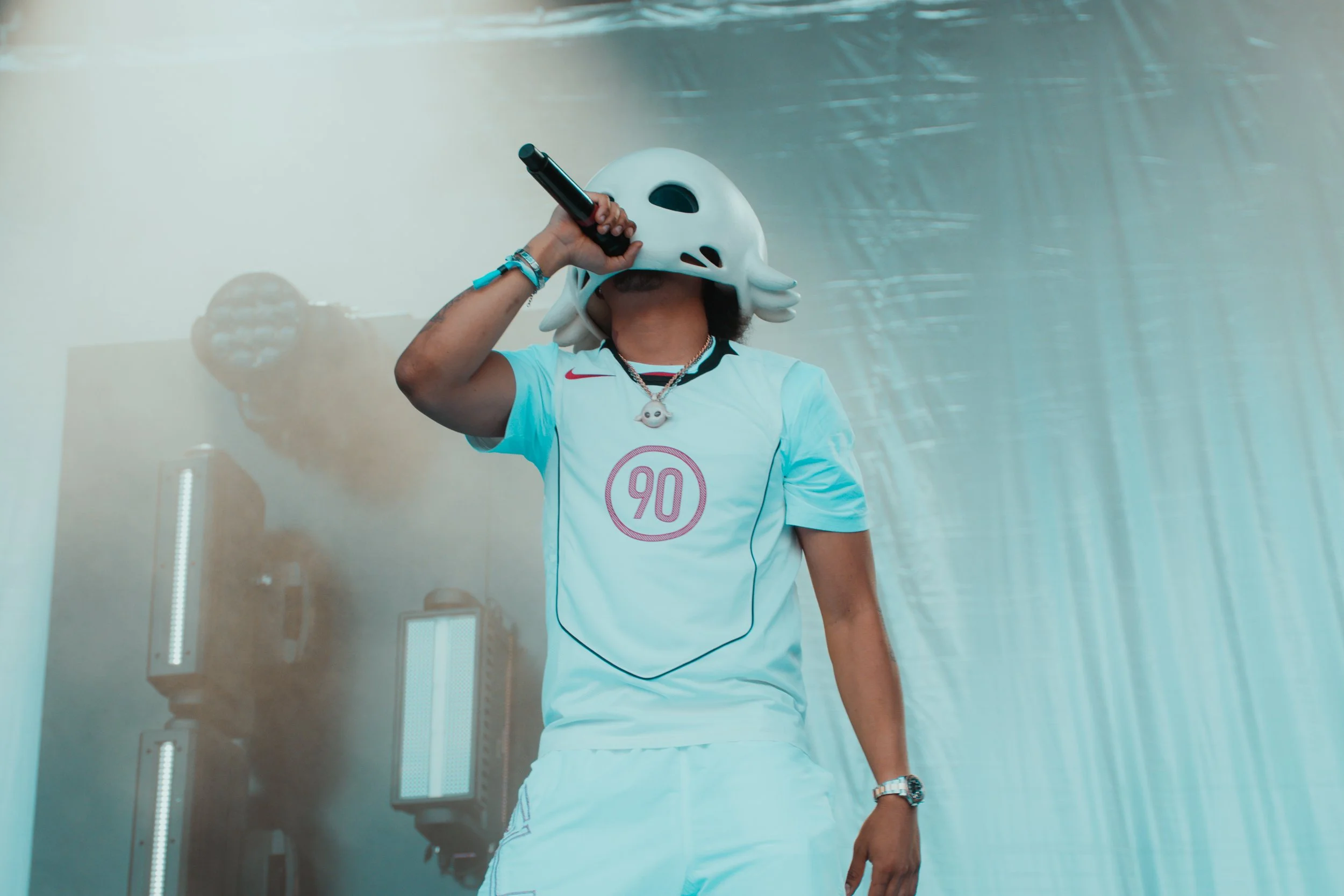 Une personne portant un casque blanc en forme de créature marine, tenant un microphone, portant un maillot de football blanc avec le chiffre 90, et des accessoires comme une chaîne avec un pendentif