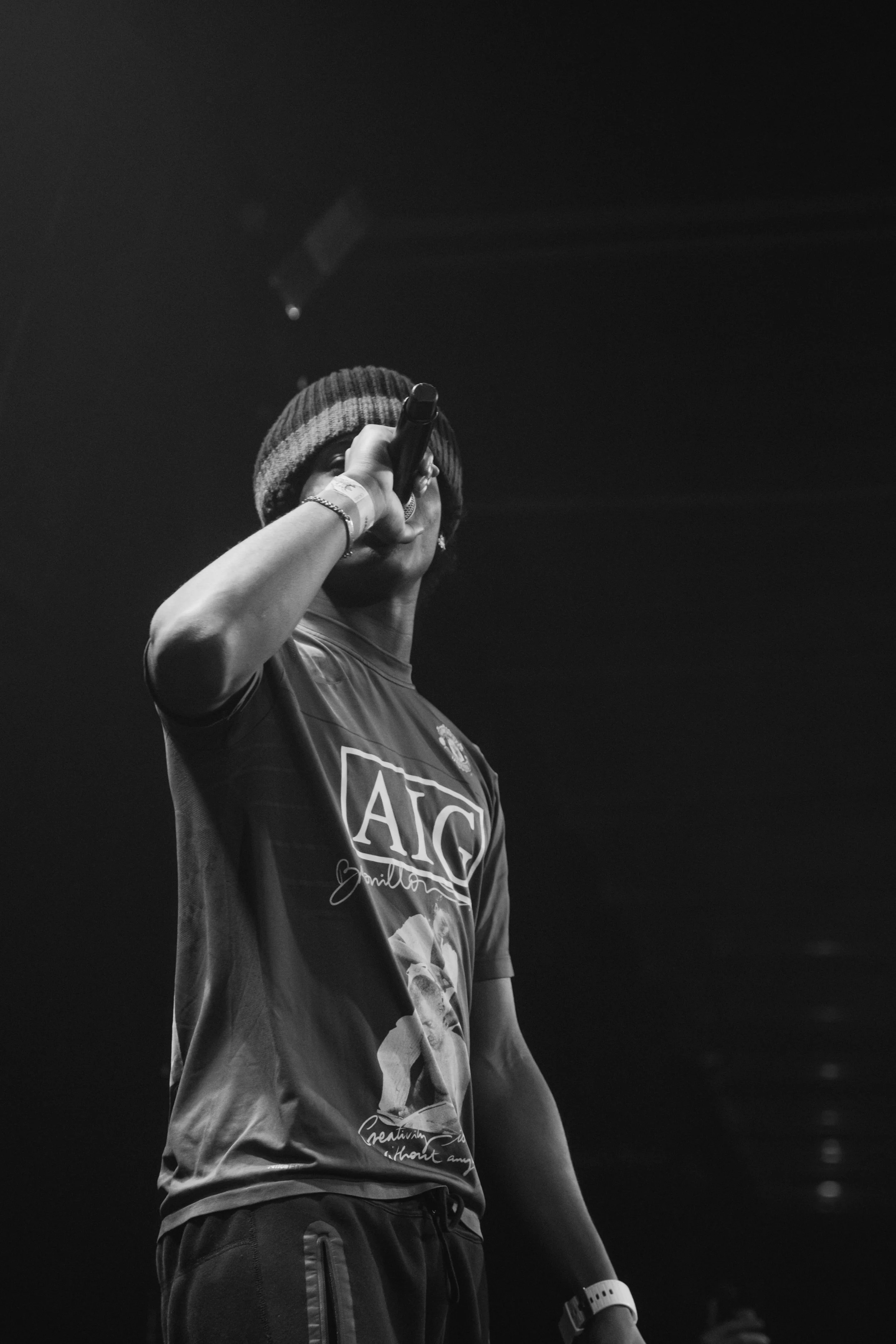 Une personne portant un bonnet de laine et un t-shirt avec un logo, tenant un microphone en train de chanter ou parler sur scène, en noir et blanc.