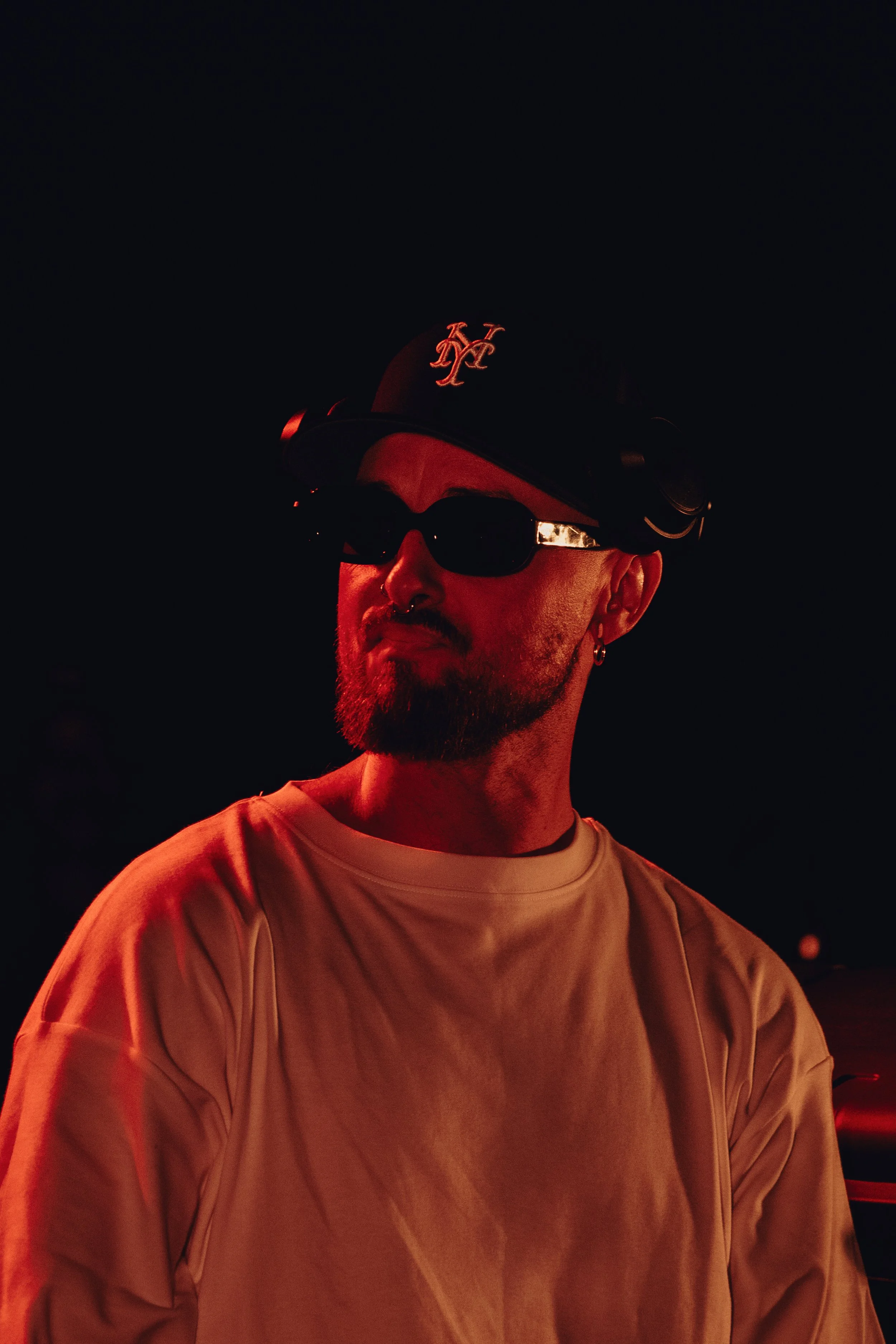 Un homme portant un chapeau noir avec le logo 'Kappa', des lunettes de soleil noires, un piercing au nez, et un t-shirt beige. Il est dans l'obscurité avec un éclairage rouge.