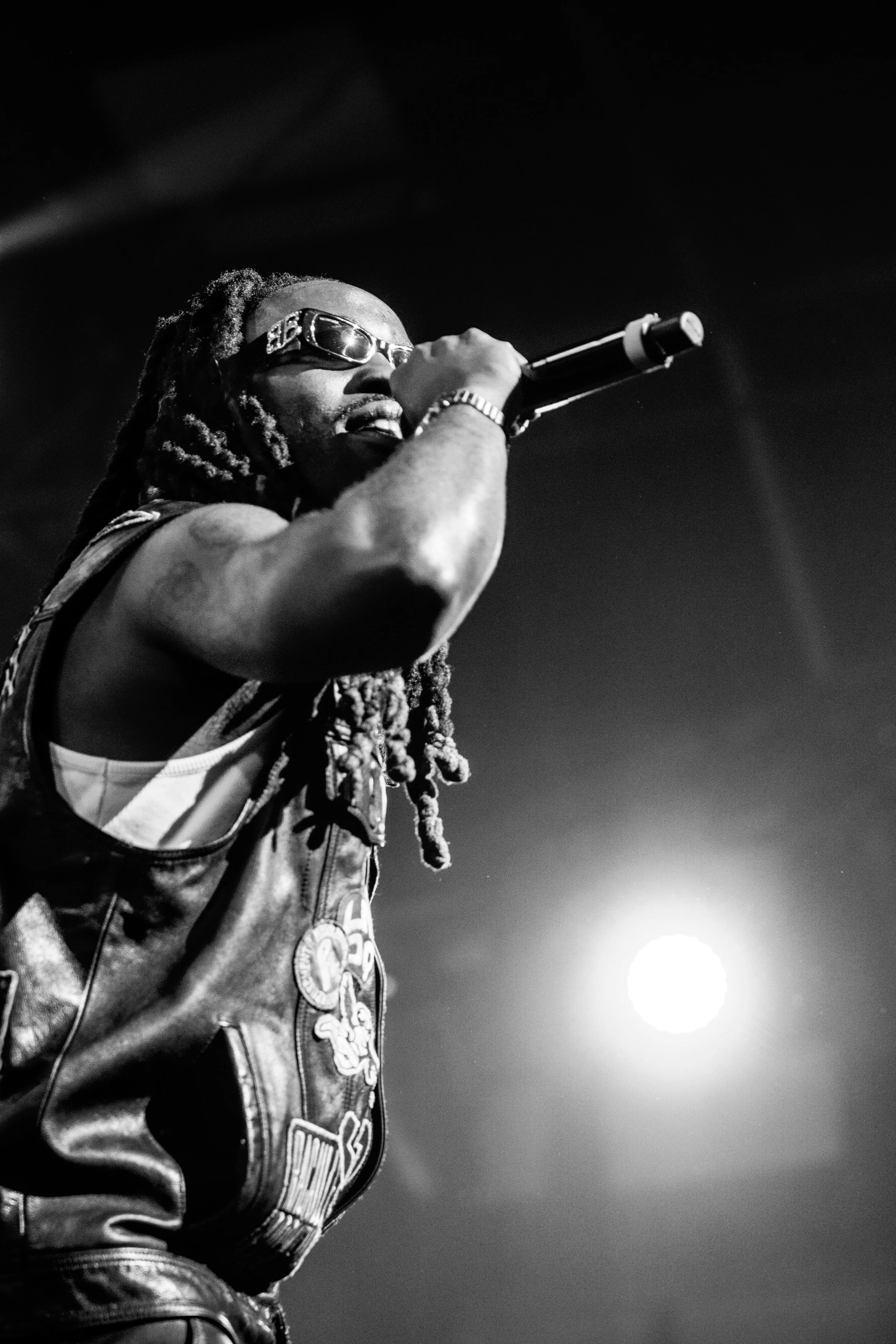 Un homme avec des dreadlocks portant des lunettes de soleil, tenant un microphone et chantant ou parlant sur scène, avec un éclairage en arrière-plan.