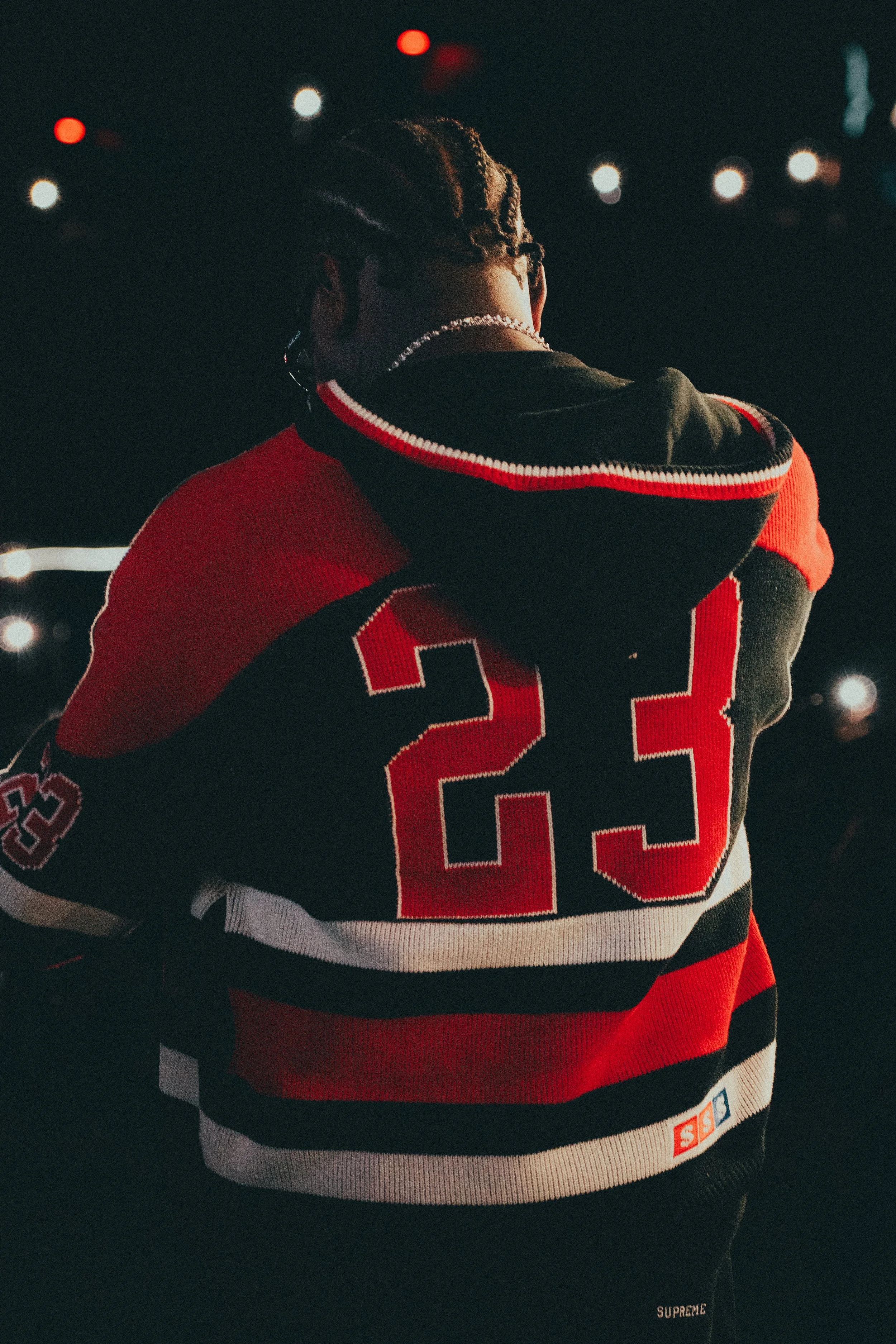 Personne vue de dos portant un maillot de hockey avec le numéro 23, dans un environnement nocturne avec des lumières floues en arrière-plan.