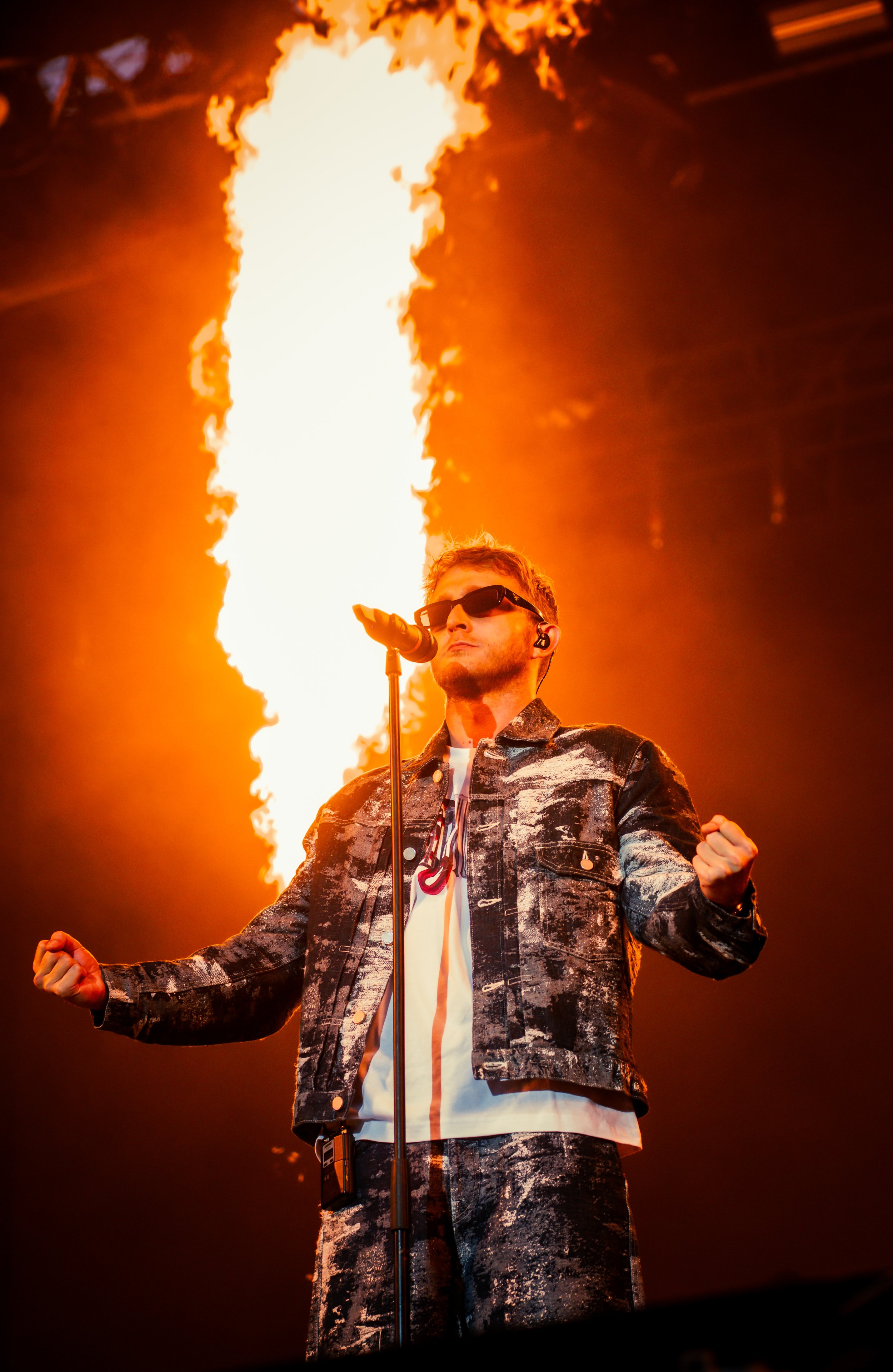 Un chanteur portant des lunettes de soleil et un vêtement camouflage se tient devant un microphone avec un feu d'artifice en arrière-plan lors d'un spectacle.
