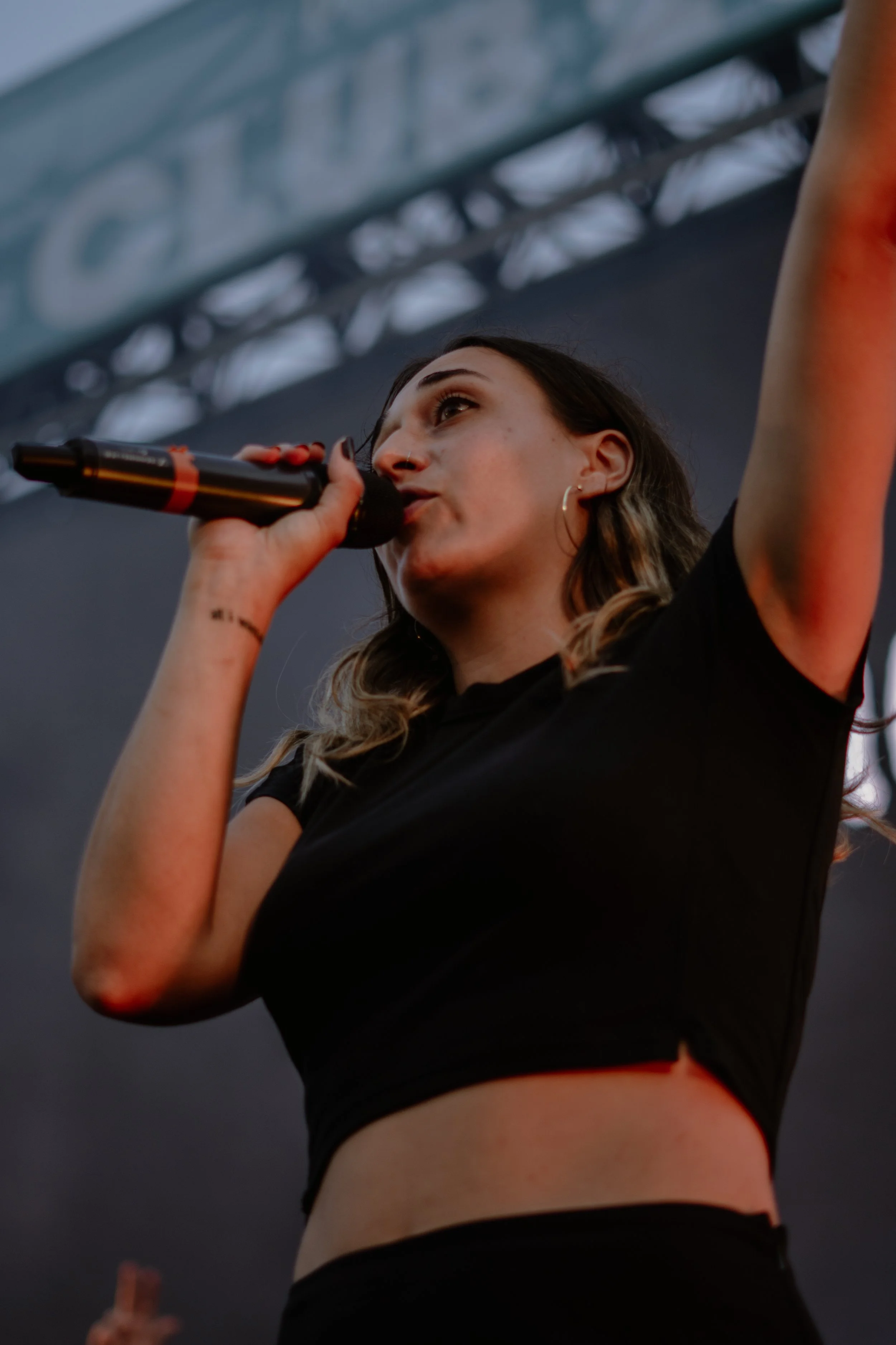 Une jeune femme chante dans un micro lors d'un concert, portant un t-shirt noir, avec un bras levé, et une scène en arrière-plan.