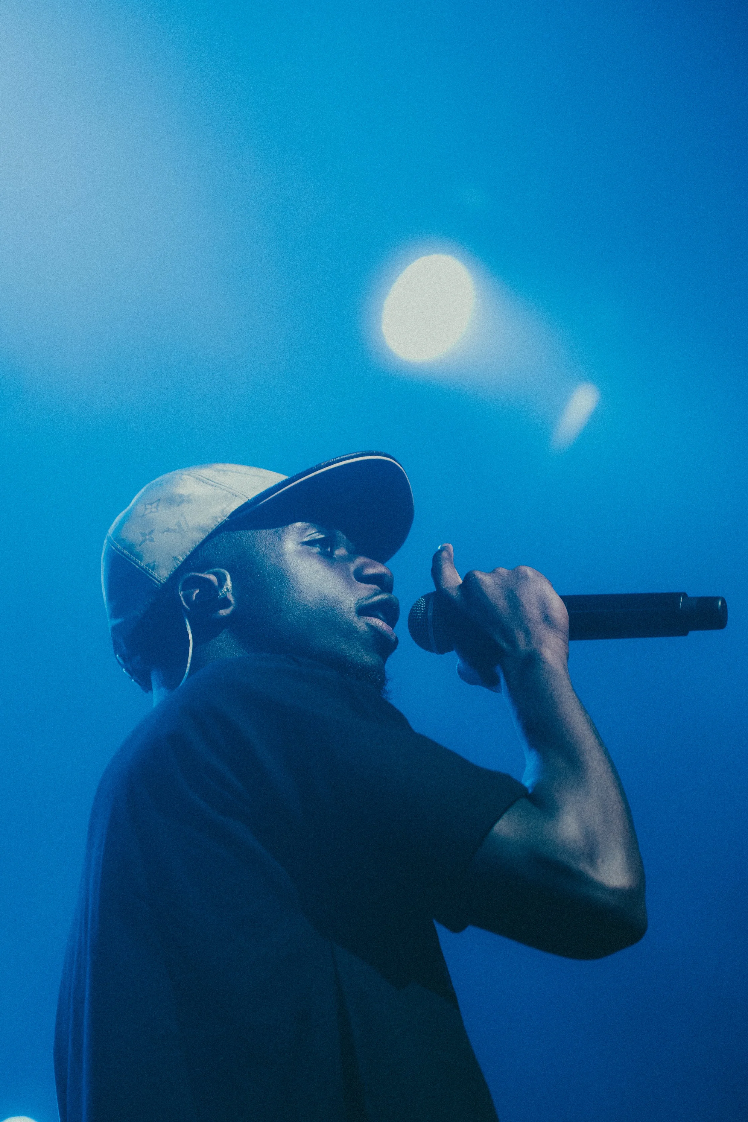 Un homme noir portant une casquette et tenant un microphone, en train de chanter ou parler sur scène, sous un éclairage bleu.