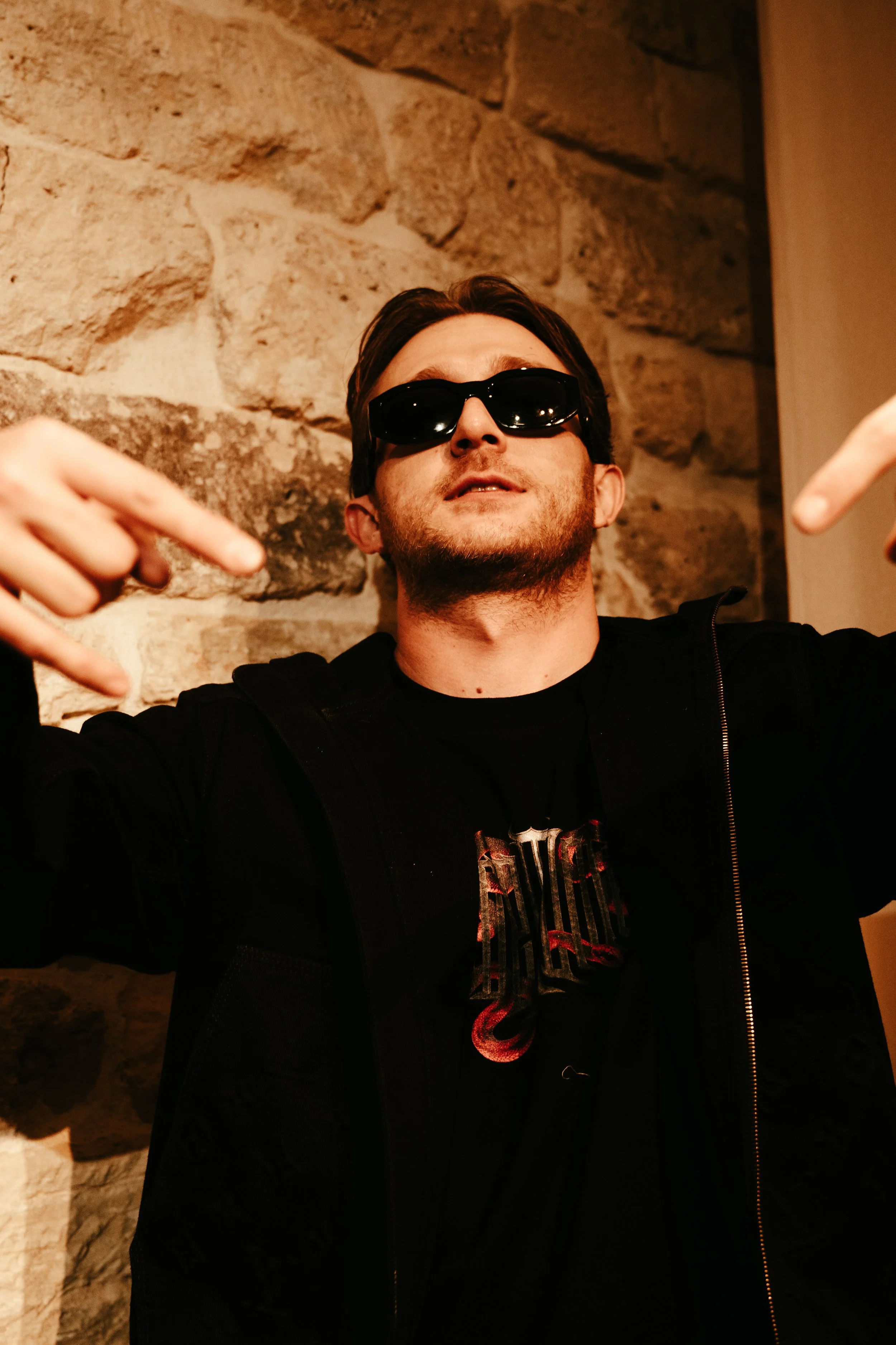 Un homme portant des lunettes de soleil noires, une veste noire et un t-shirt noir avec un logo rouge, posant devant un mur en pierre, faisant un geste de main avec les doigts.