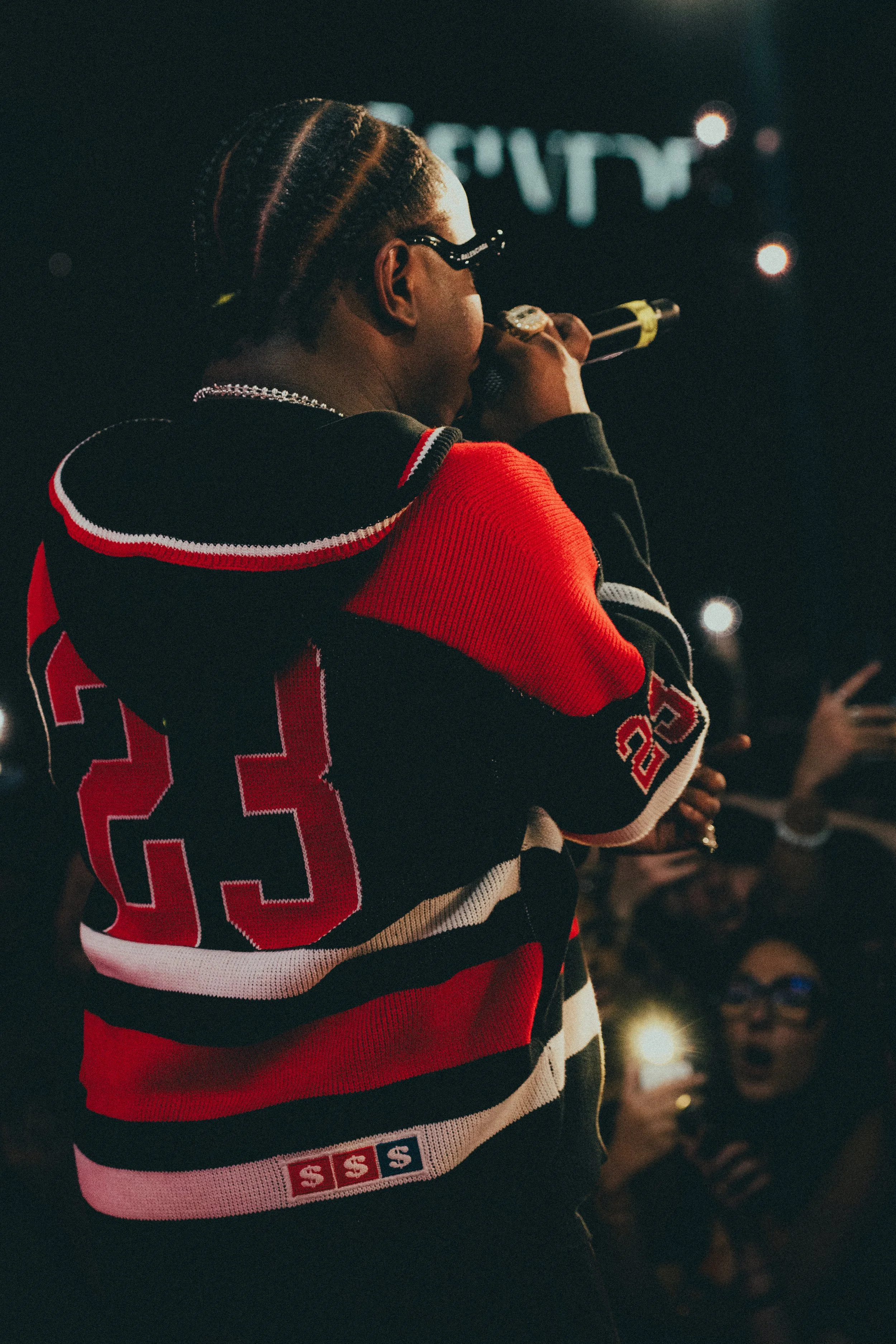 Un rappeur chante sur scène lors d'un concert, portant un sweat à gros logo rouge, noir et blanc, avec des gens dans le fond prenant des photos avec leur téléphone.
