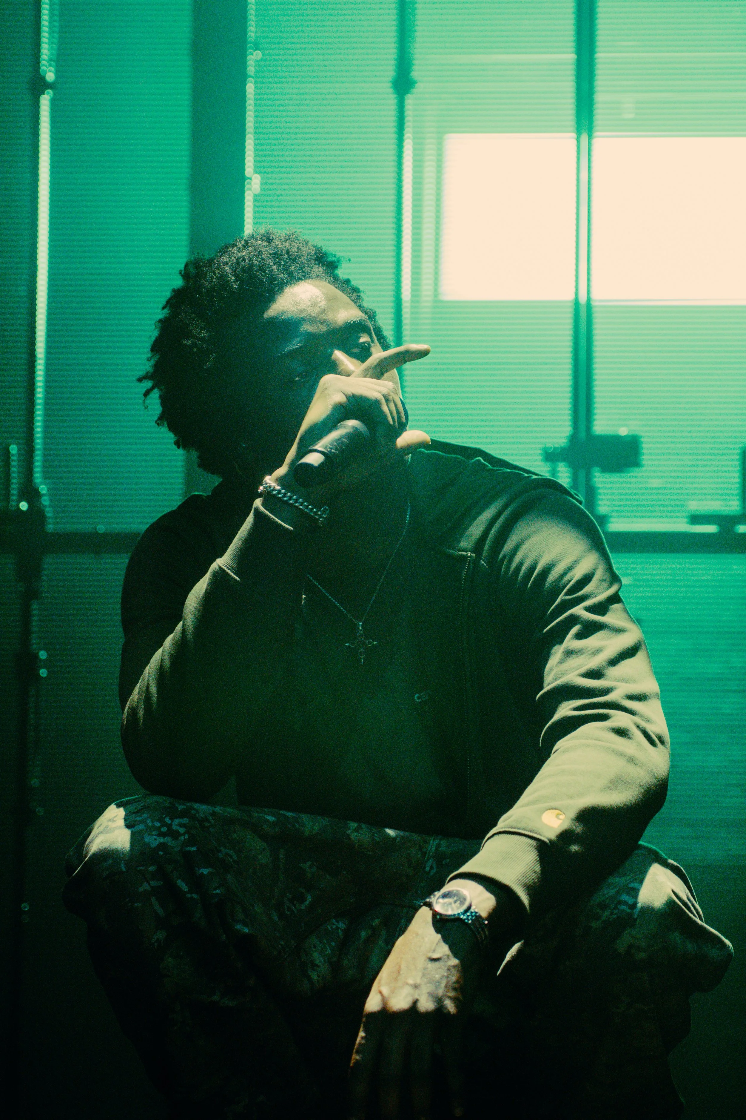 Jeune homme assis en position décontractée, tenant un microphone, avec des fenêtres en arrière-plan.