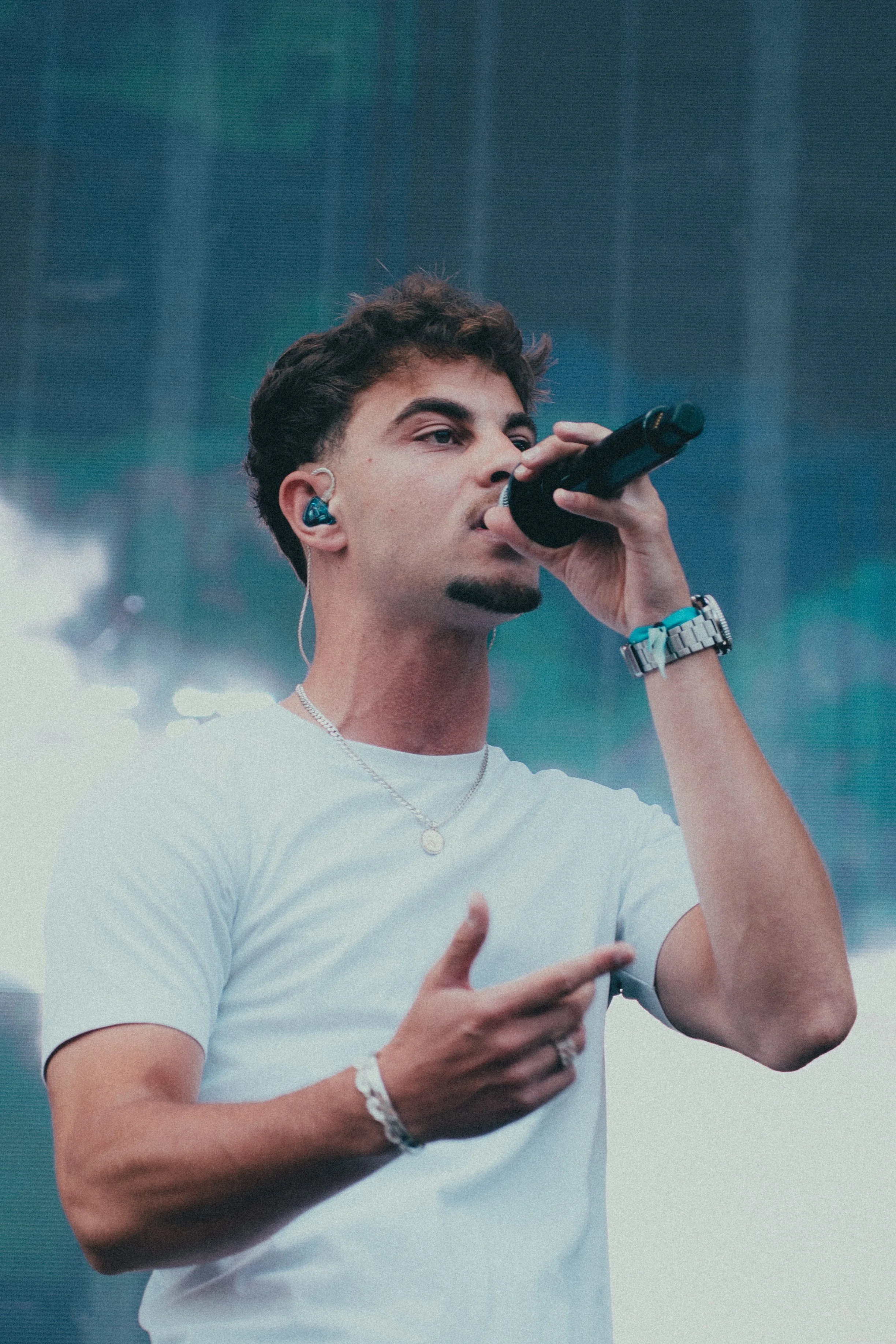 Un jeune homme chante dans un micro devant un arrière-plan flou, portant un t-shirt blanc, des écouteurs bleus et une montre.