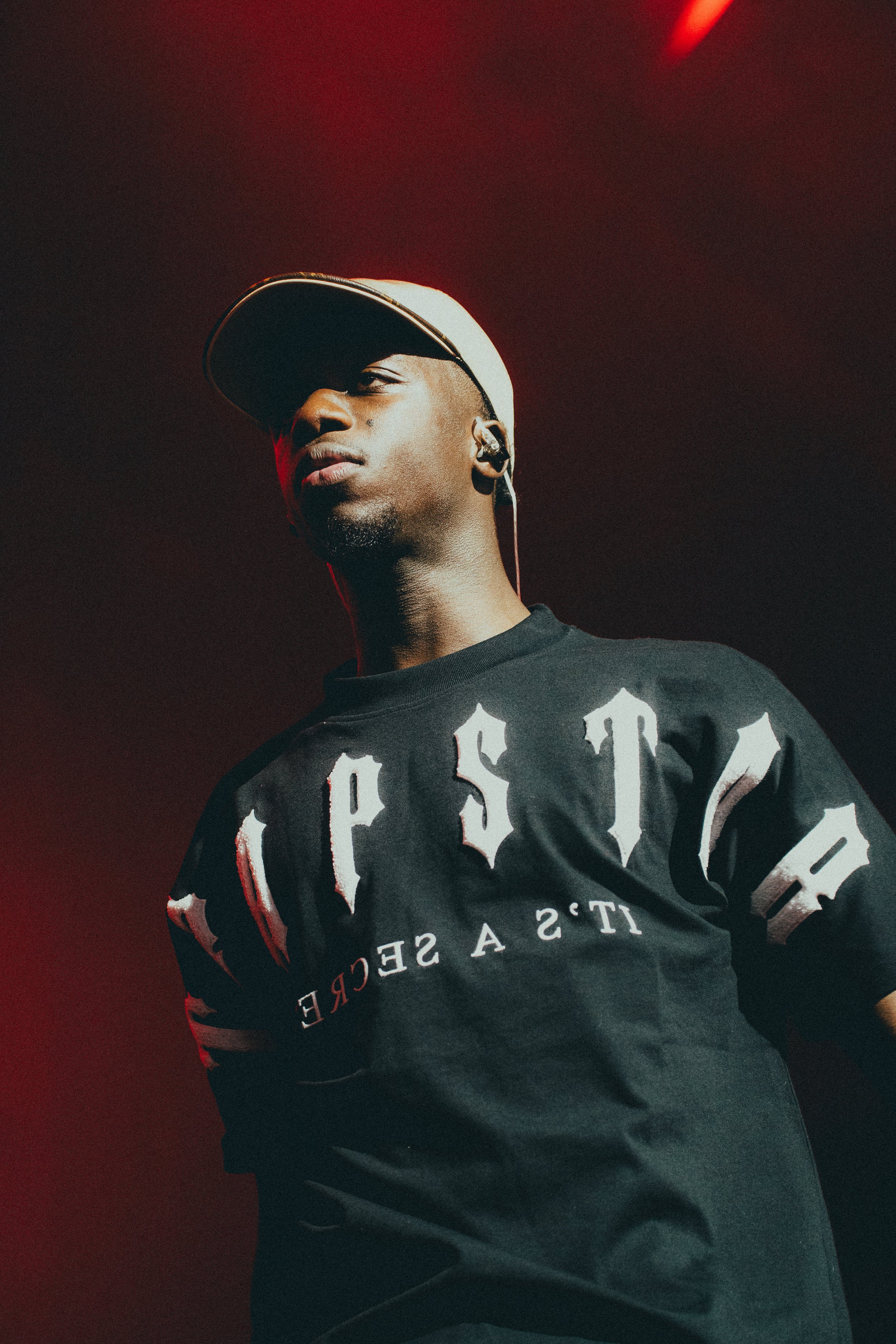 Un homme portant une casquette blanche et un t-shirt noir avec des lettres blanches et un slogan, en train de se produire sur scène avec un éclairage rouge en arrière-plan.
