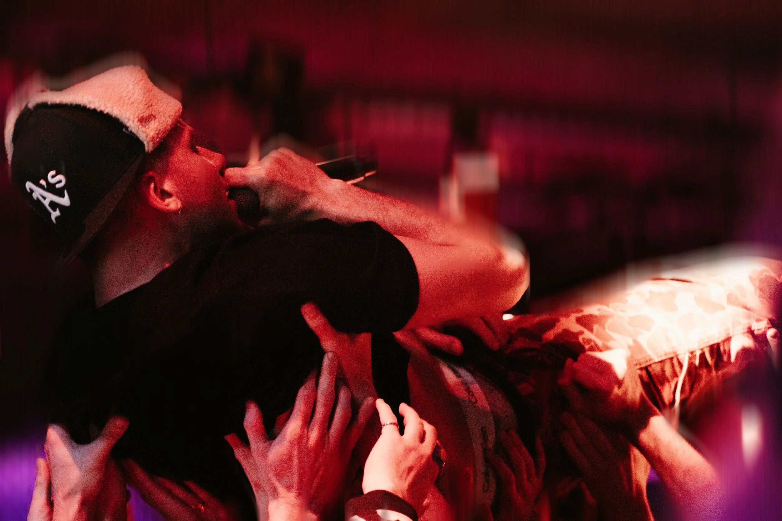 Un jeune homme portant un chapeau noir avec un t-shirt noir, tenant un microphone et chantant sur scène, entouré de plusieurs mains qui le soutiennent, dans un environnement obscur avec un éclairage rouge.
