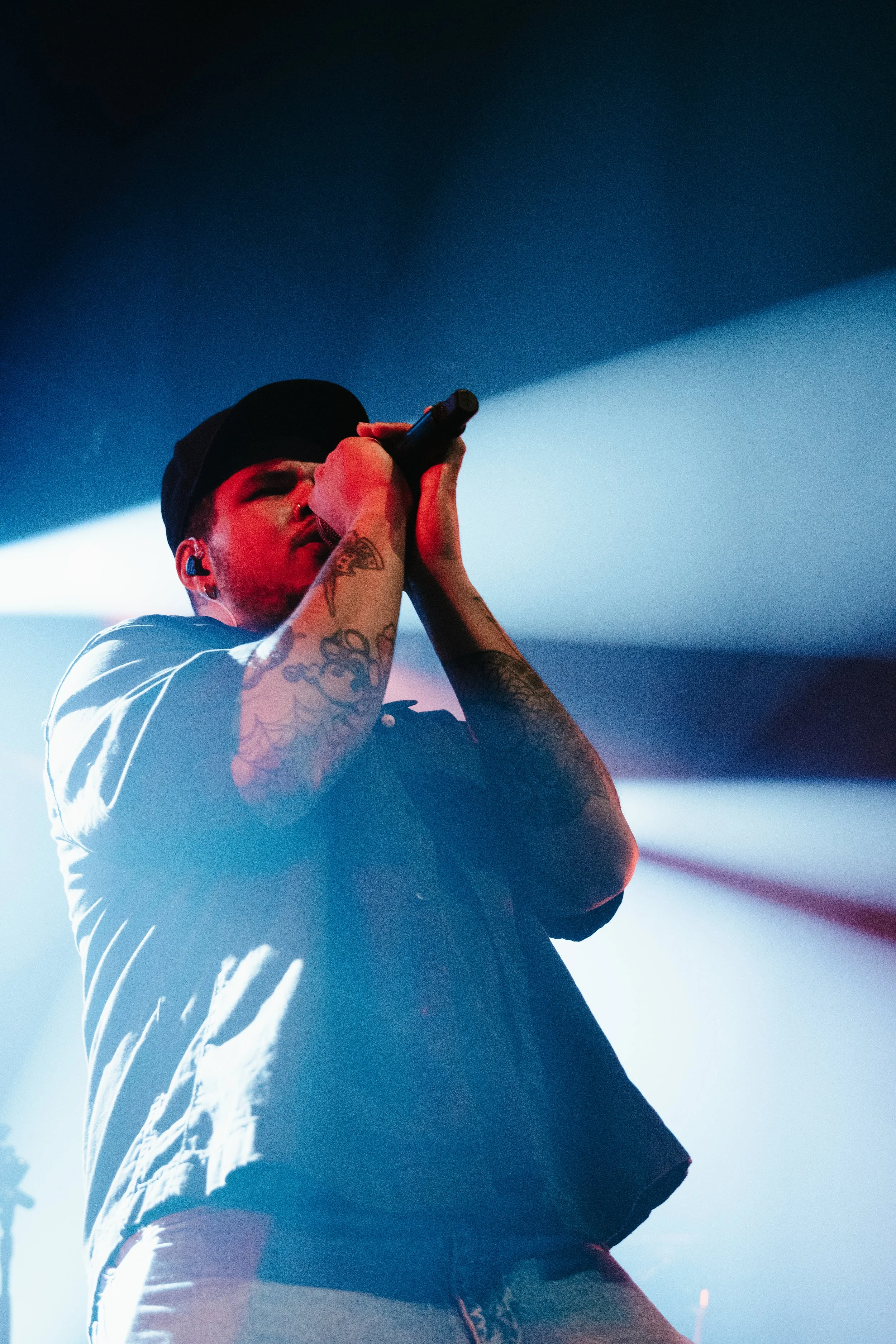 Un chanteur avec des tatouages sur les bras, portant une casquette noire, chante dans un microphone sur scène avec un éclairage de scène coloré.