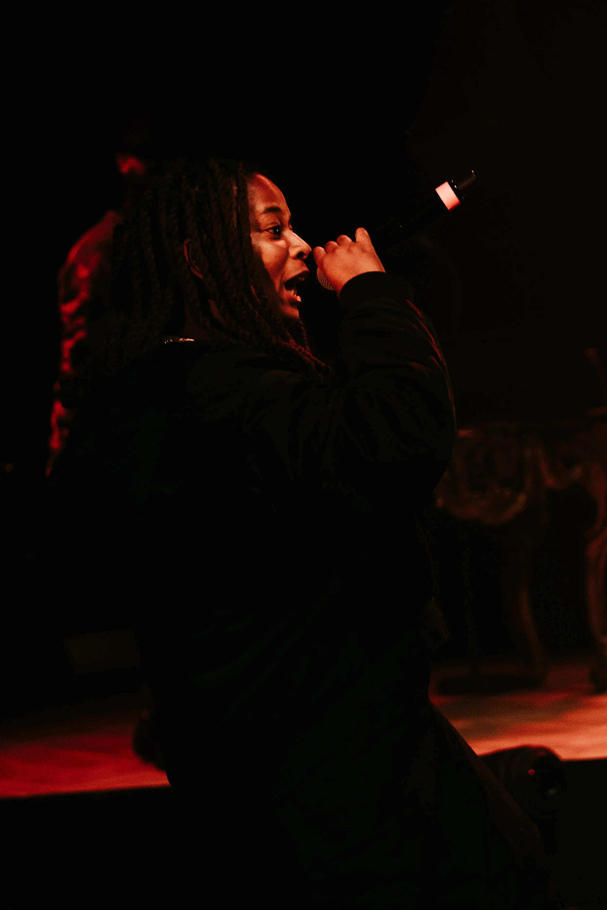 Une femme chante en tenant un microphone sur scène, avec un éclairage rougeâtre dans un environnement sombre.