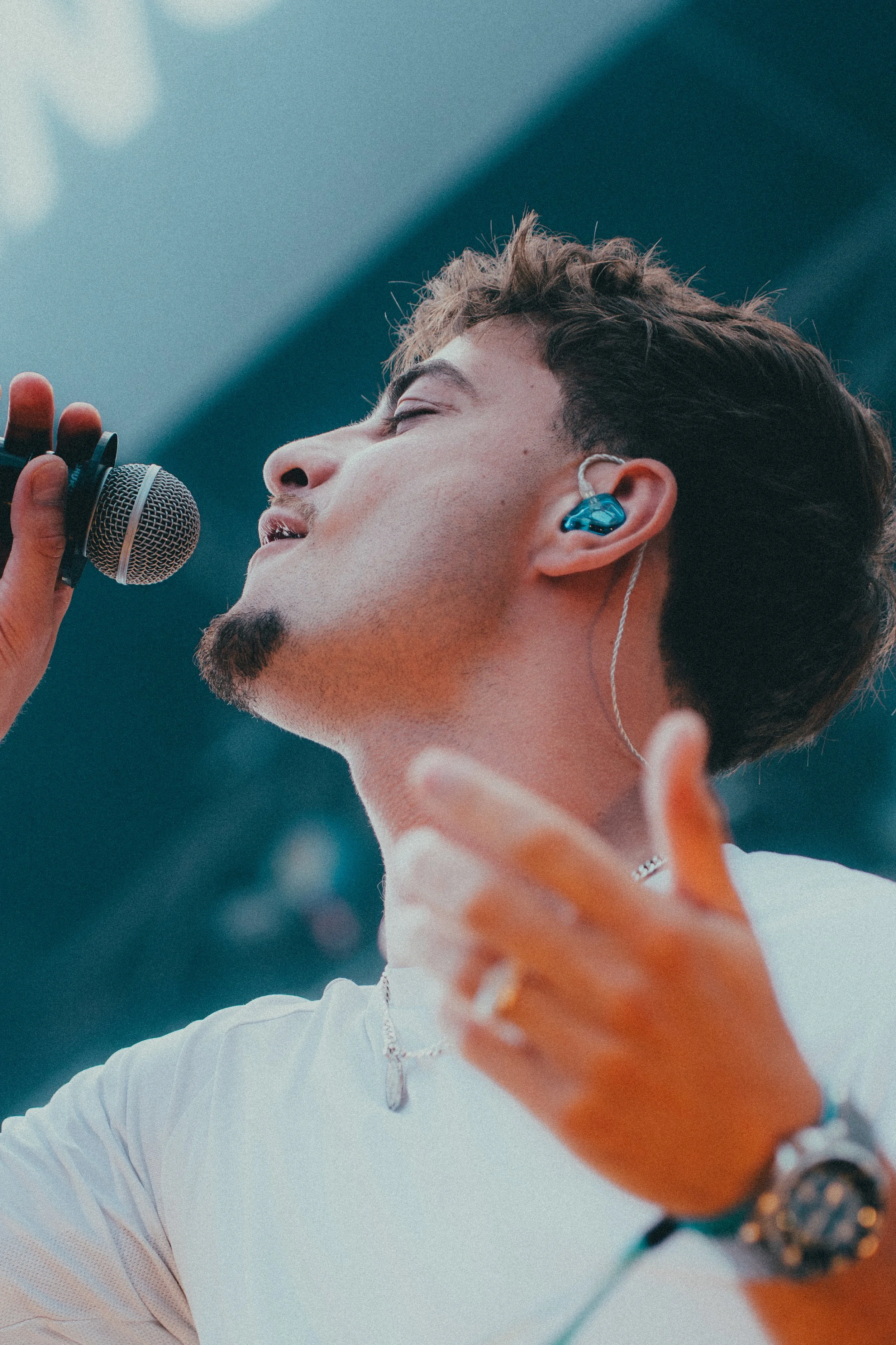 Un homme chante avec un micro, portant un écouteur bleu et une montre, en costume blanc.