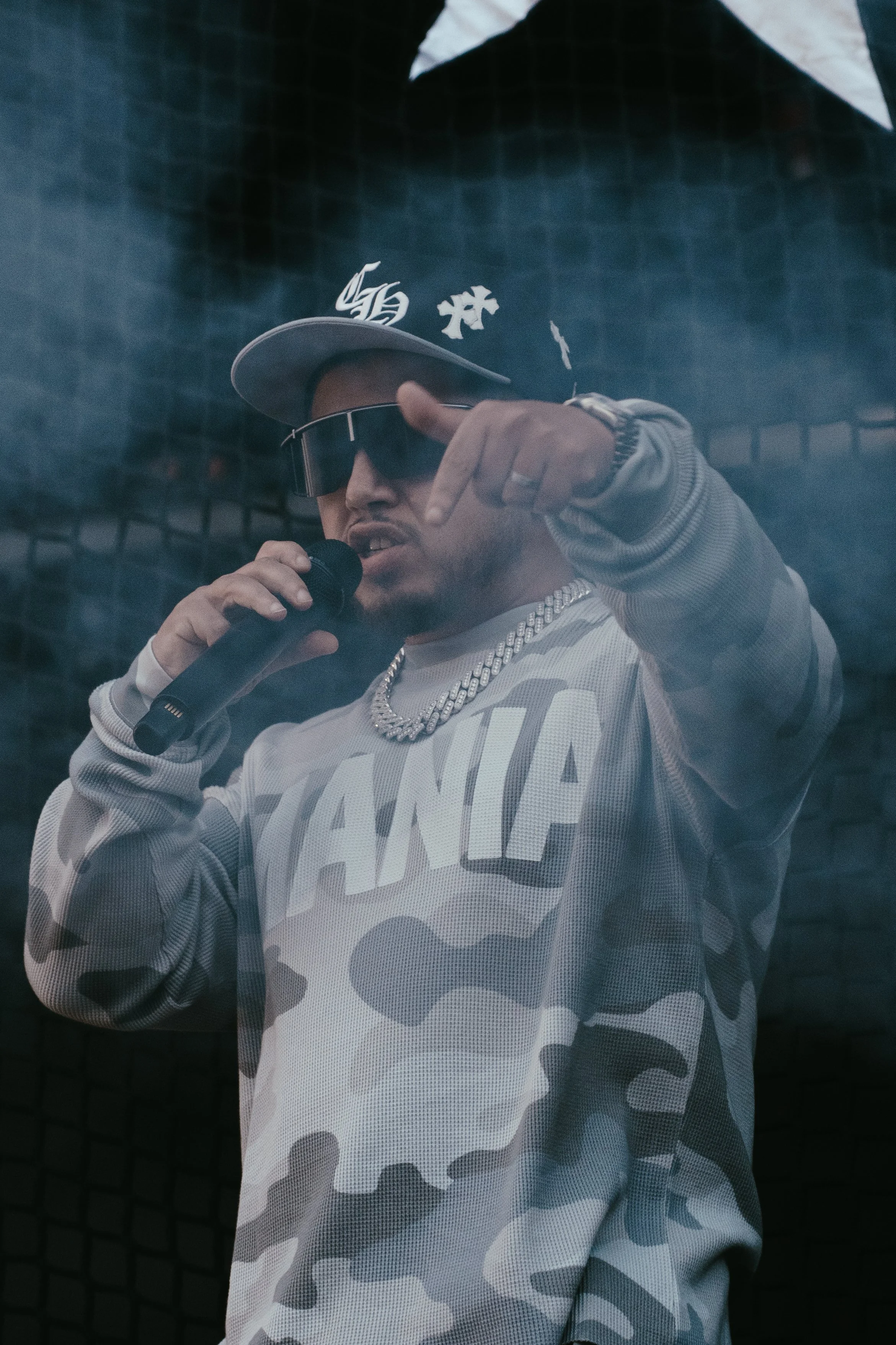 Un homme portant une casquette, des lunettes de soleil, un sweat à motifs camouflage et une chaîne en or, tenant un microphone et faisant un geste de la main.