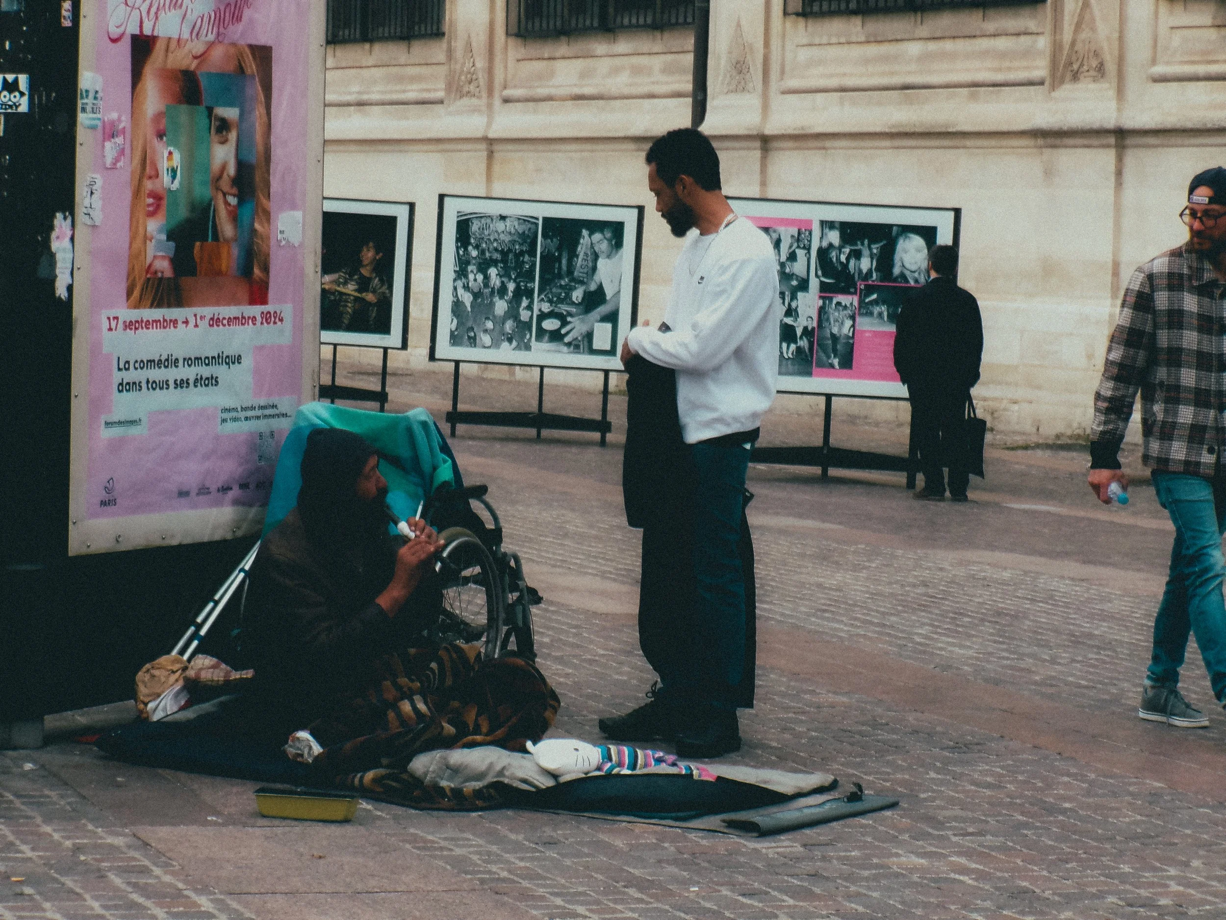 Une femme sans-abri assise par terre, mangeant un sandwich, avec un chariot à proximité, face à un homme qui la regarde.