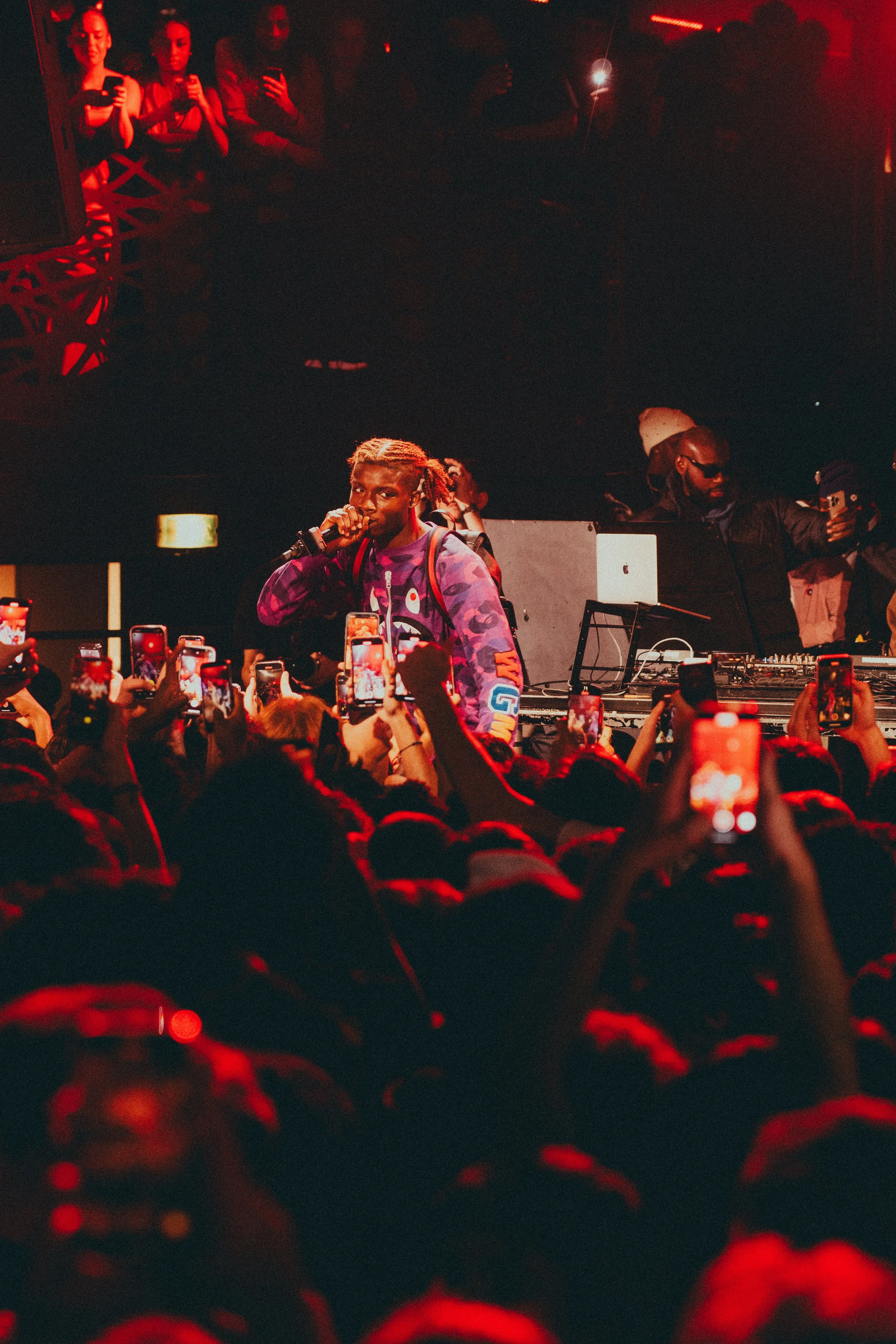 Un artiste performe lors d'un concert ou d'un événement musical, entouré d'une foule nombreuse qui filme la scène avec leurs téléphones. La scène est éclairée en rouge, créant une ambiance intense et énergique.
