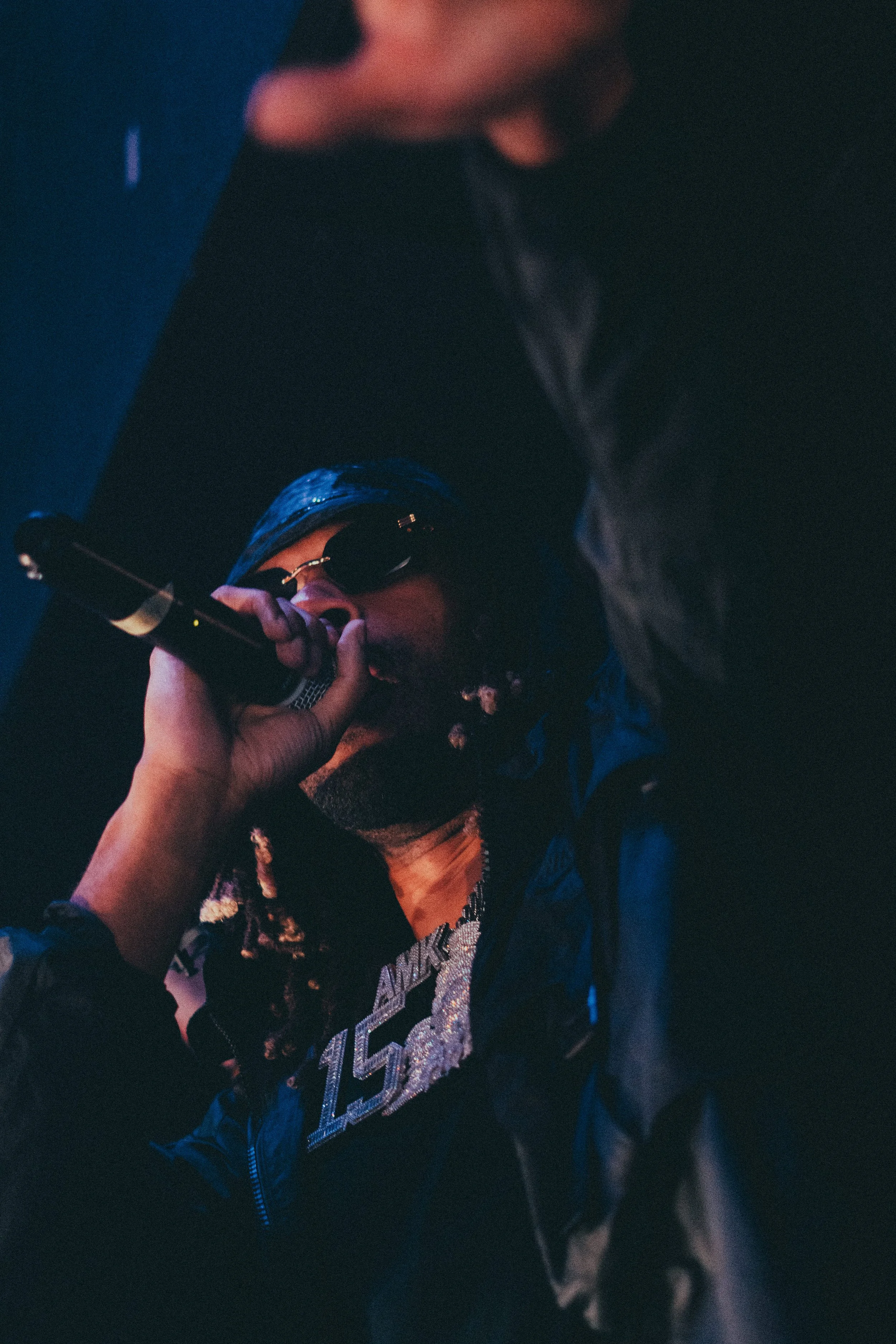 Un rappeur portant des lunettes de soleil et un chapeau, tenant un microphone en train de performer sur scène dans une atmosphère sombre et colorée.