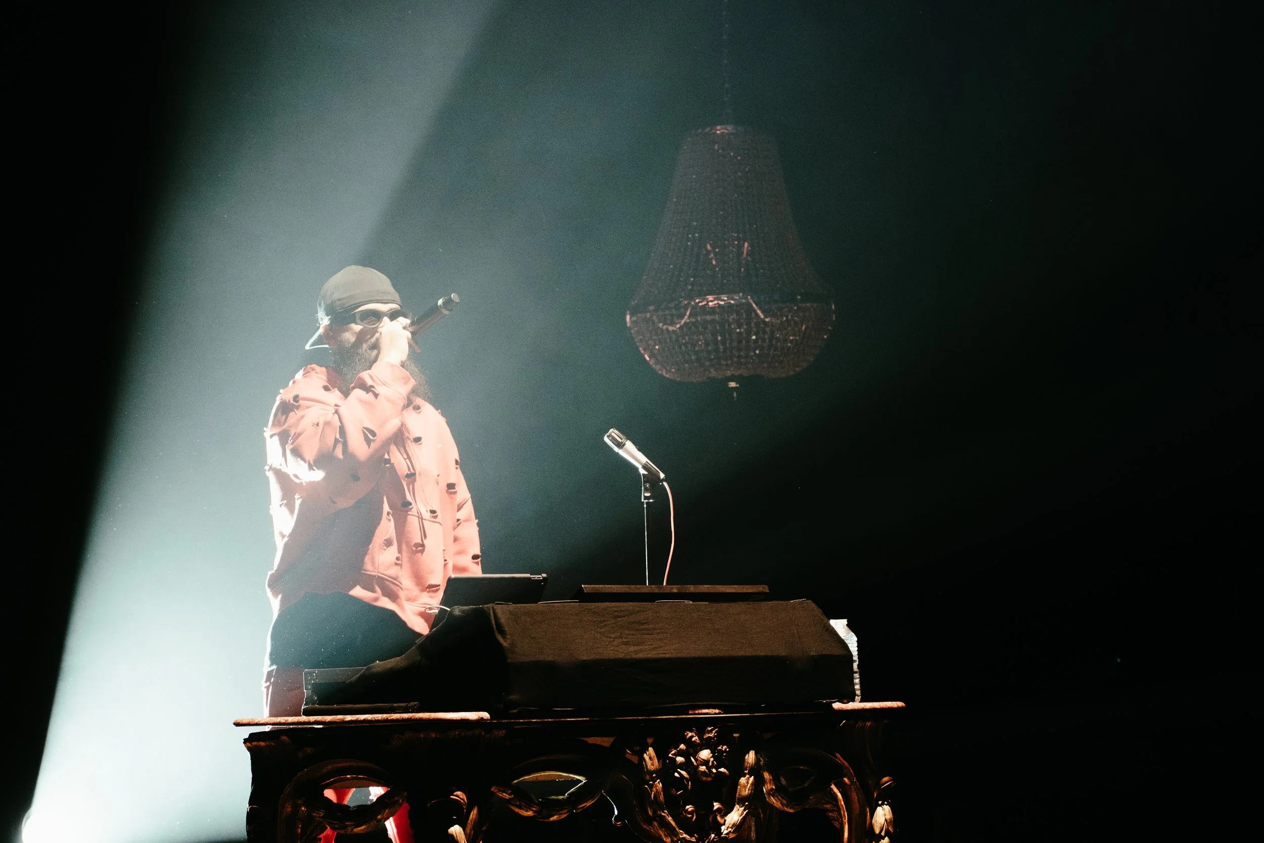 Un homme avec un chapeau et une barbe chante dans un microphone, éclairé par un faisceau lumineux, derrière lui une lampe suspendue en cristal, et il se tient devant un pupitre en bois orné de sculptures.