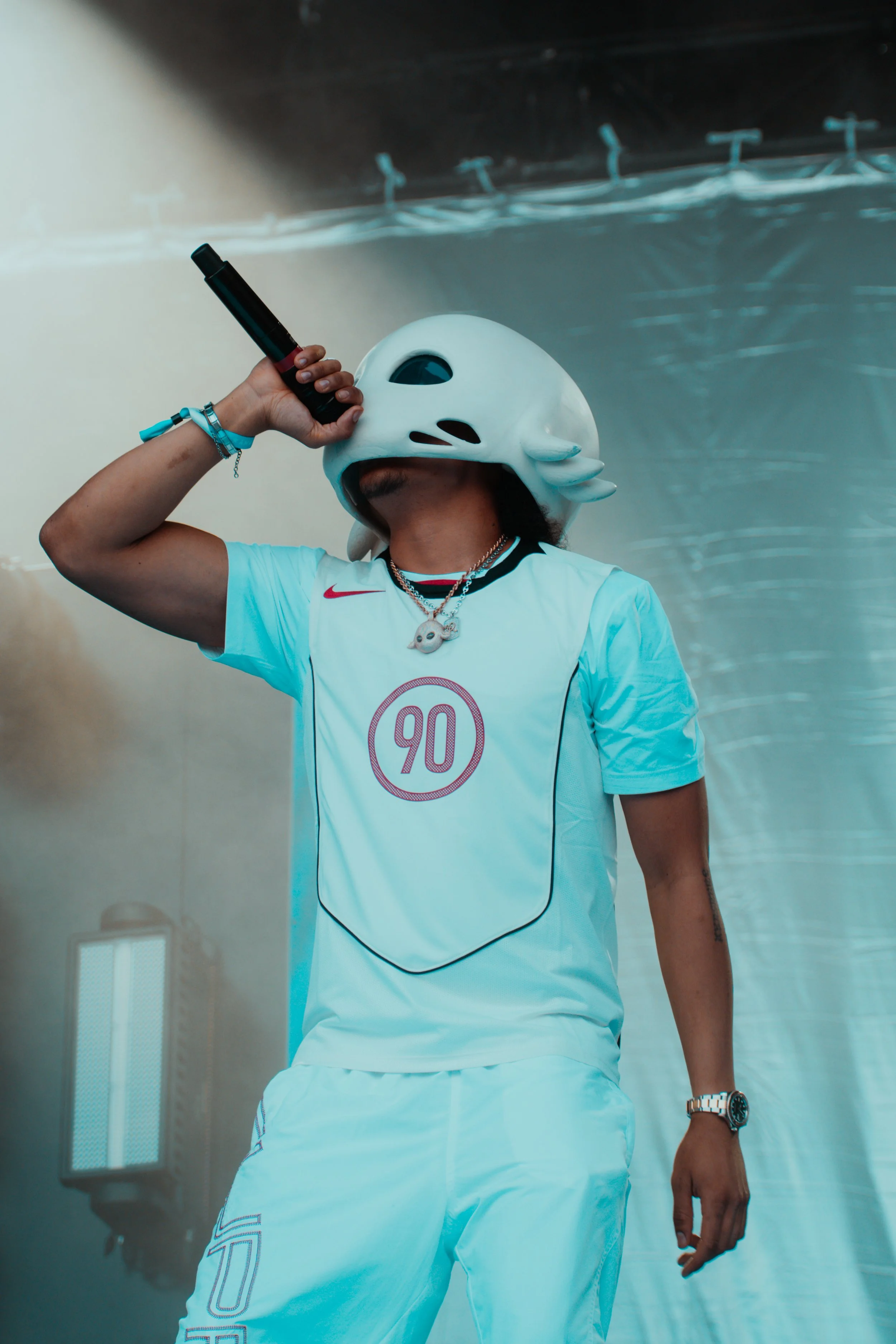 Une personne porte un casque de style animal et une tenue de sport blanche avec le numéro 90, tenant un microphone et faisant un geste de performance.
