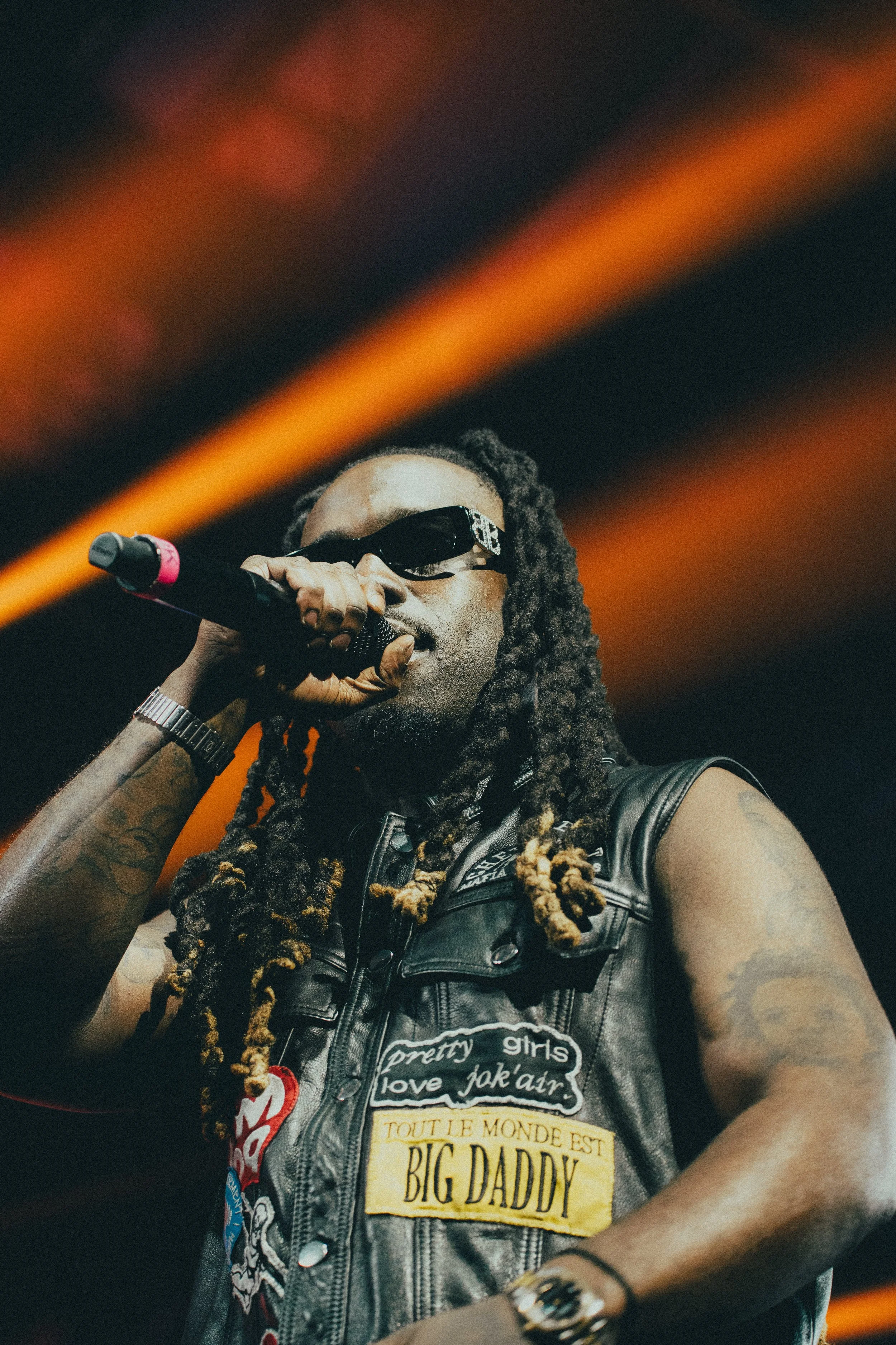 Un homme avec des dreadlocks, porteur de lunettes de soleil, tient un microphone lors d'une performance sur scène, portant une veste en cuir avec des patches.