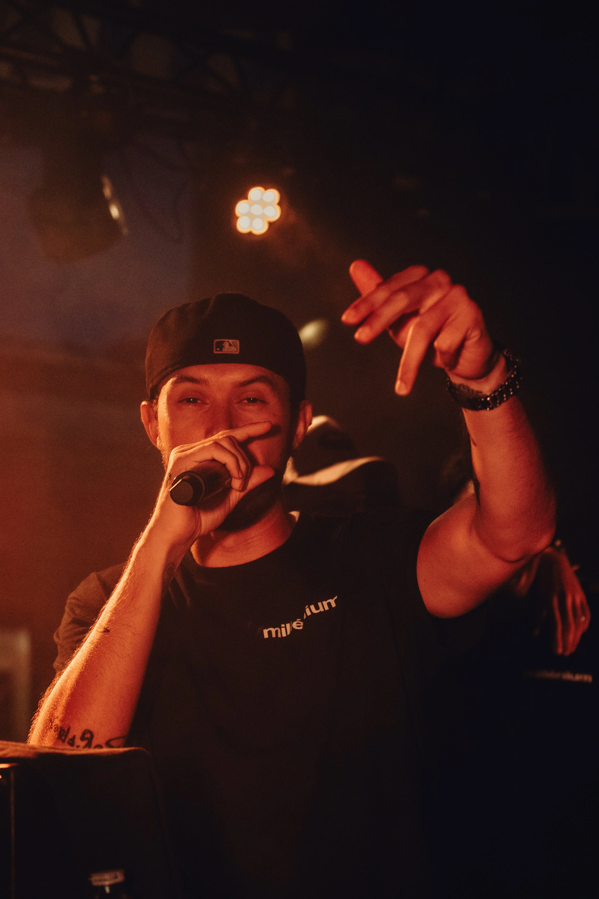 Un homme portant une casquette noire, tenant un microphone, parlant ou chantant dans une scène sombre, avec des lumières en arrière-plan et un bras tatoué visible.