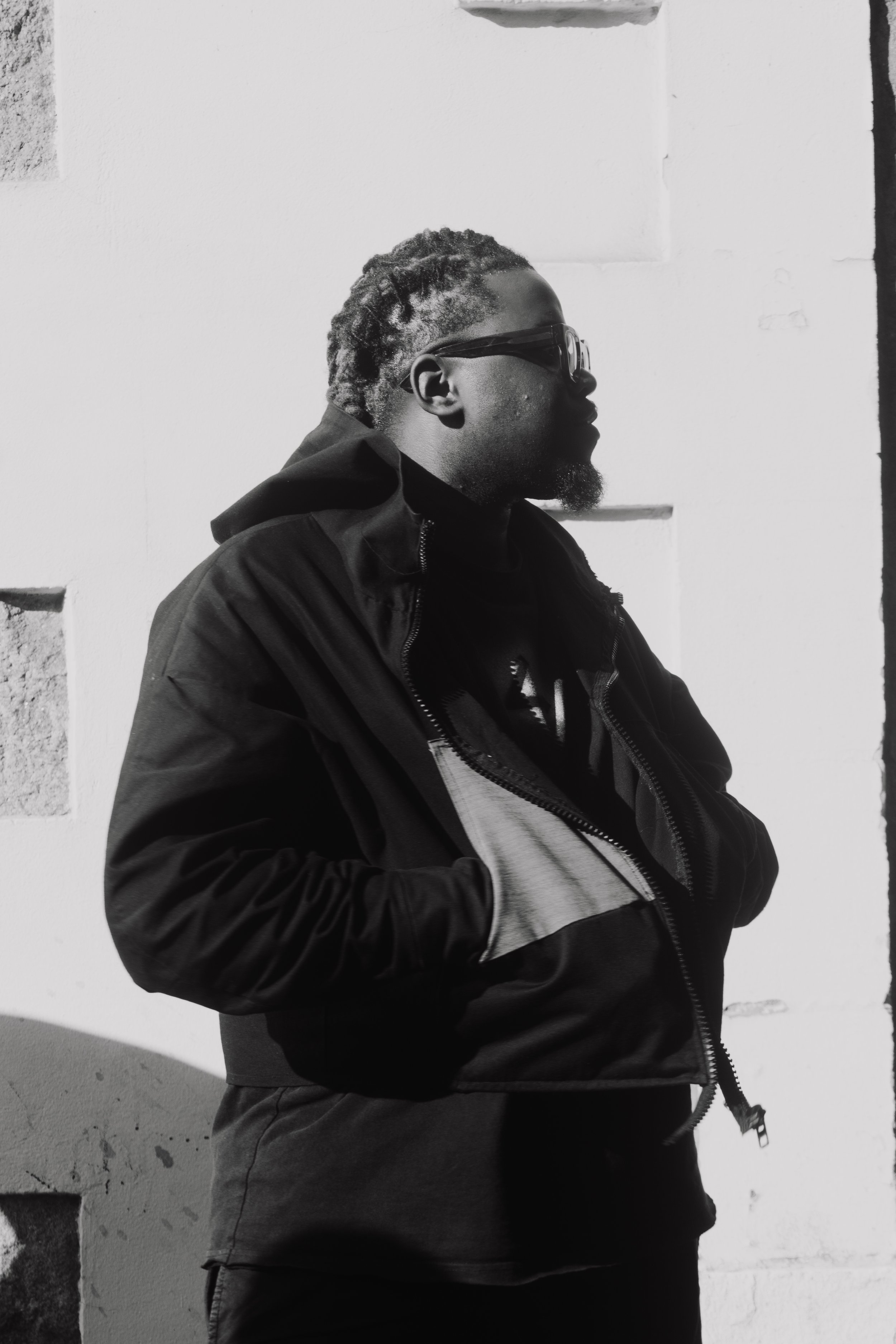 Un homme portant des lunettes de soleil et une veste sombre, regardant vers la droite, en contre-jour, à côté d'un mur blanc.