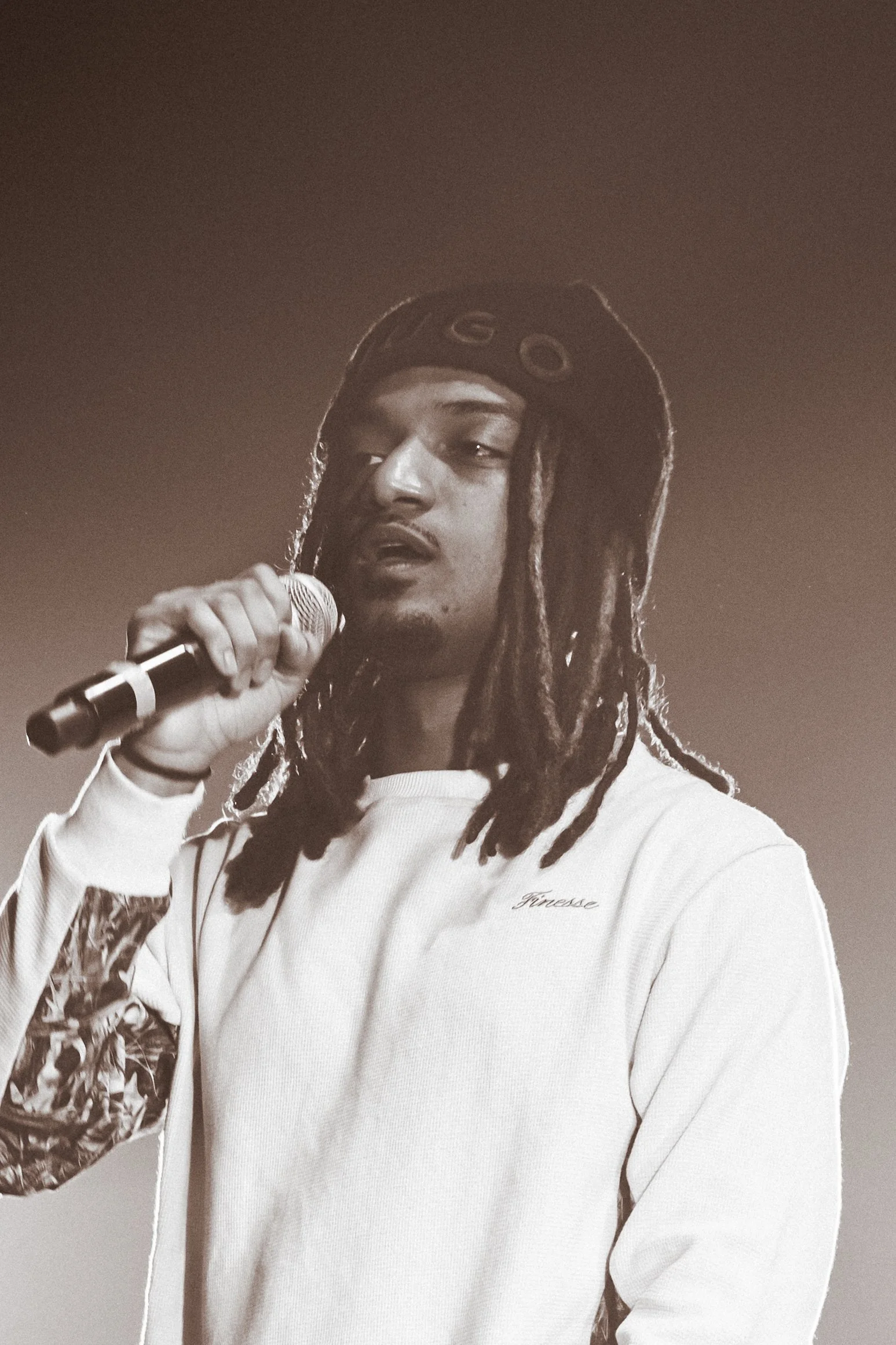 Un homme avec des dreadlocks, portant un bonnet noir avec le mot 'GATO', tient un microphone et semble chanter ou parler, avec un fond sombre.