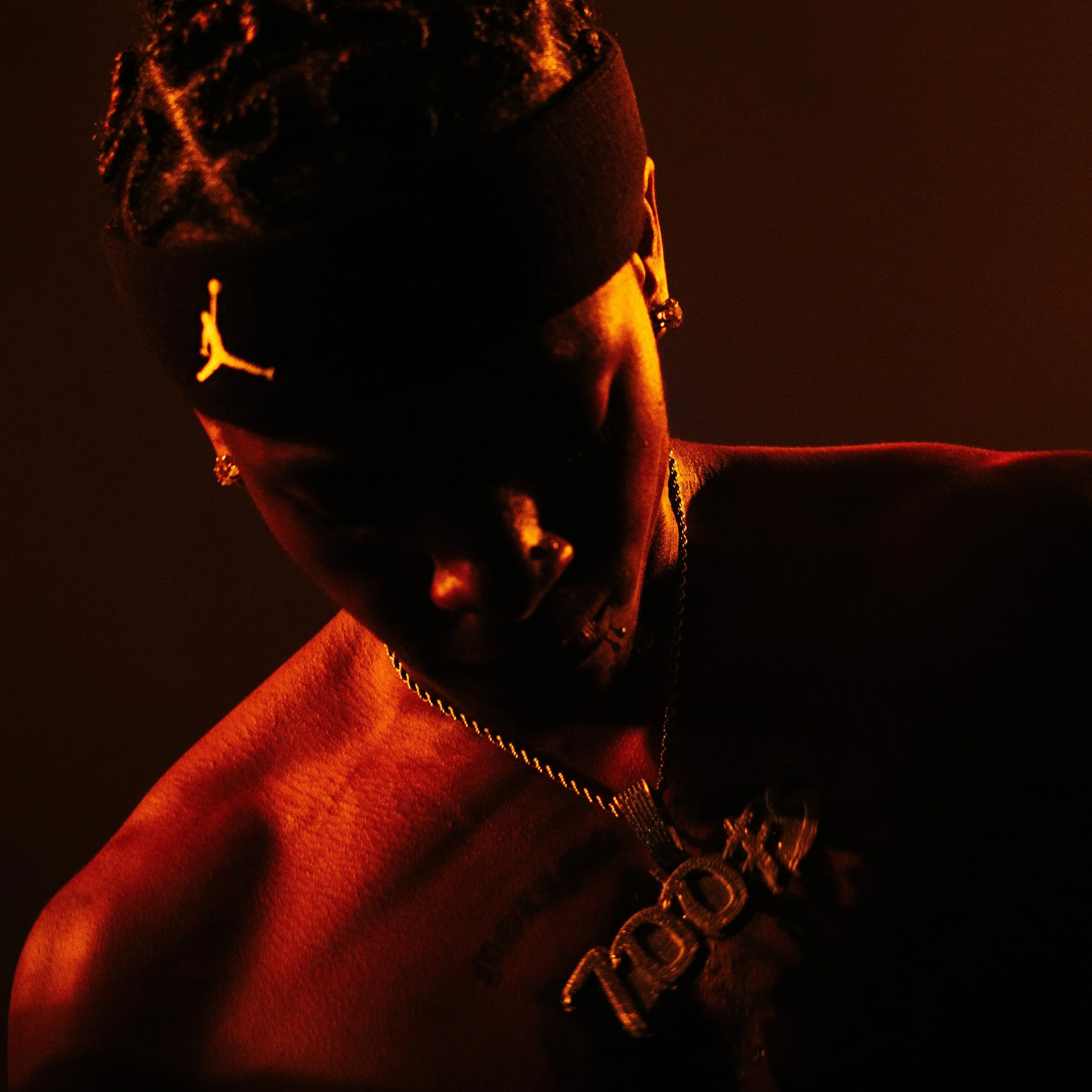 Un jeune homme porte un bonnet noir avec un logo Jumpman en blanc, un collier avec une médaille et a une tatouage sur la poitrine. La scène est sombre avec une lumière orange/rouge chaude qui illumine partiellement son visage et ses épaules.