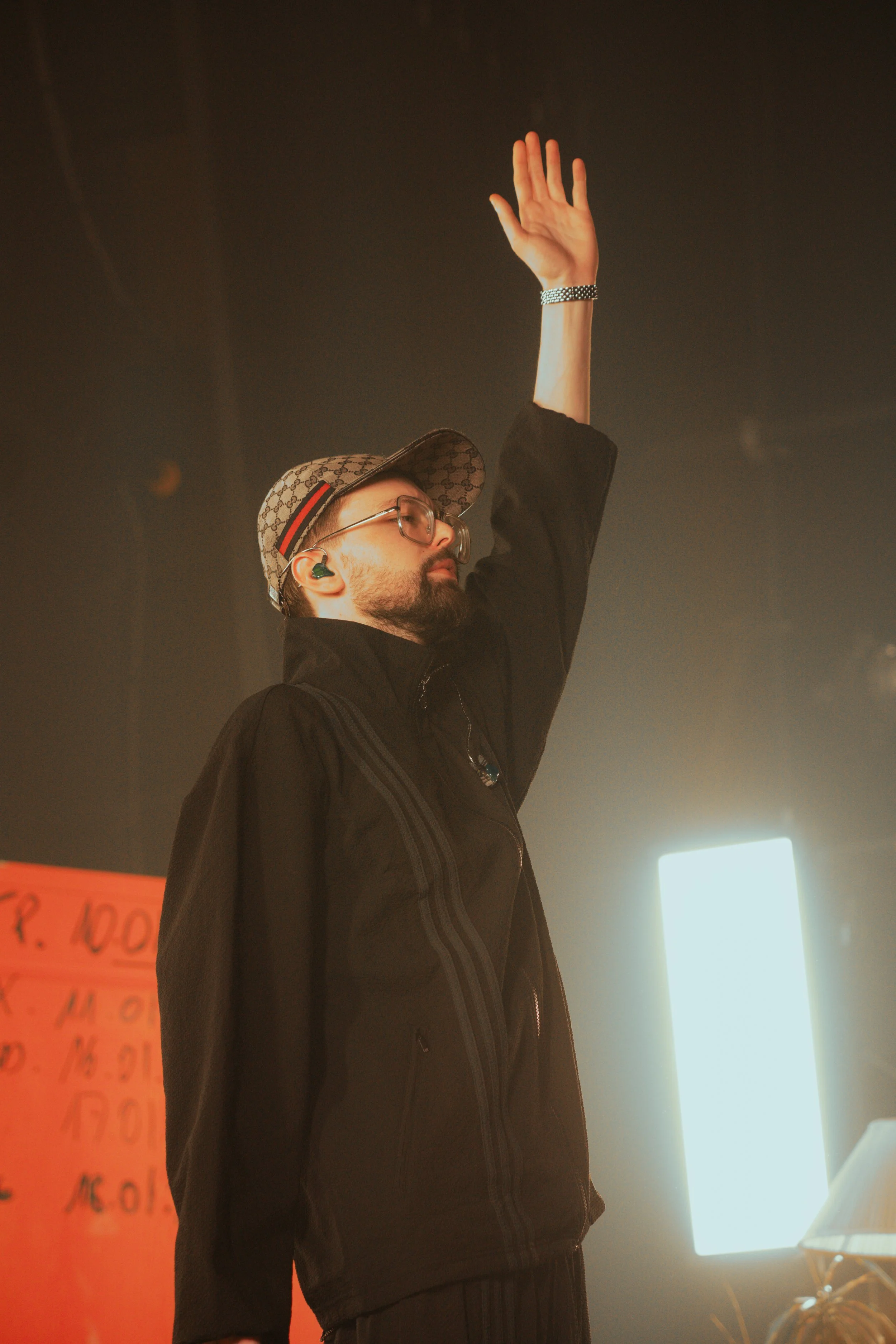 Homme avec lunettes, casquette Gucci, écouteurs verts, vêtement noir, lève la main dans une scène sombre avec une lumière forte en arrière-plan.