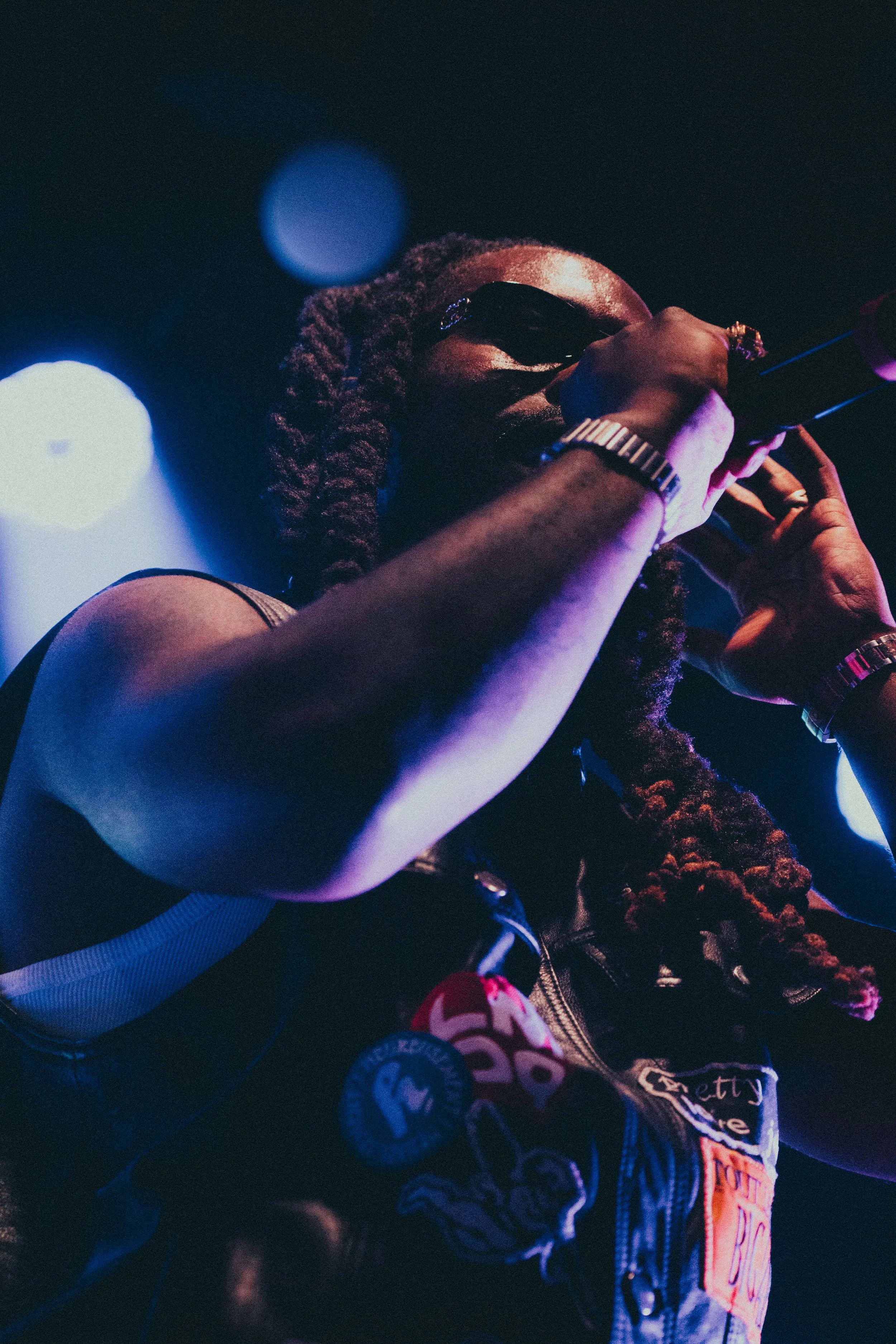 Une personne portant des lunettes de soleil, une veste en cuir avec des patchs, joue de la guitare électrique sous un éclairage coloré dans un environnement sombre.