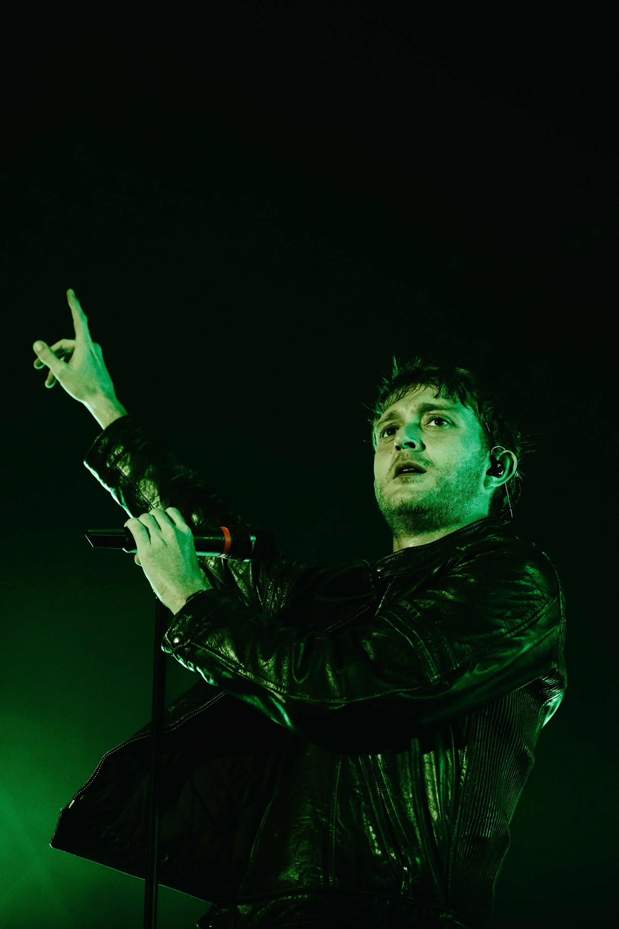 Un homme chante sur scène, portant une veste en cuir noire, avec un éclairage vert dramatique et tenant un microphone dans sa main droite, pointant le doigt vers le haut.